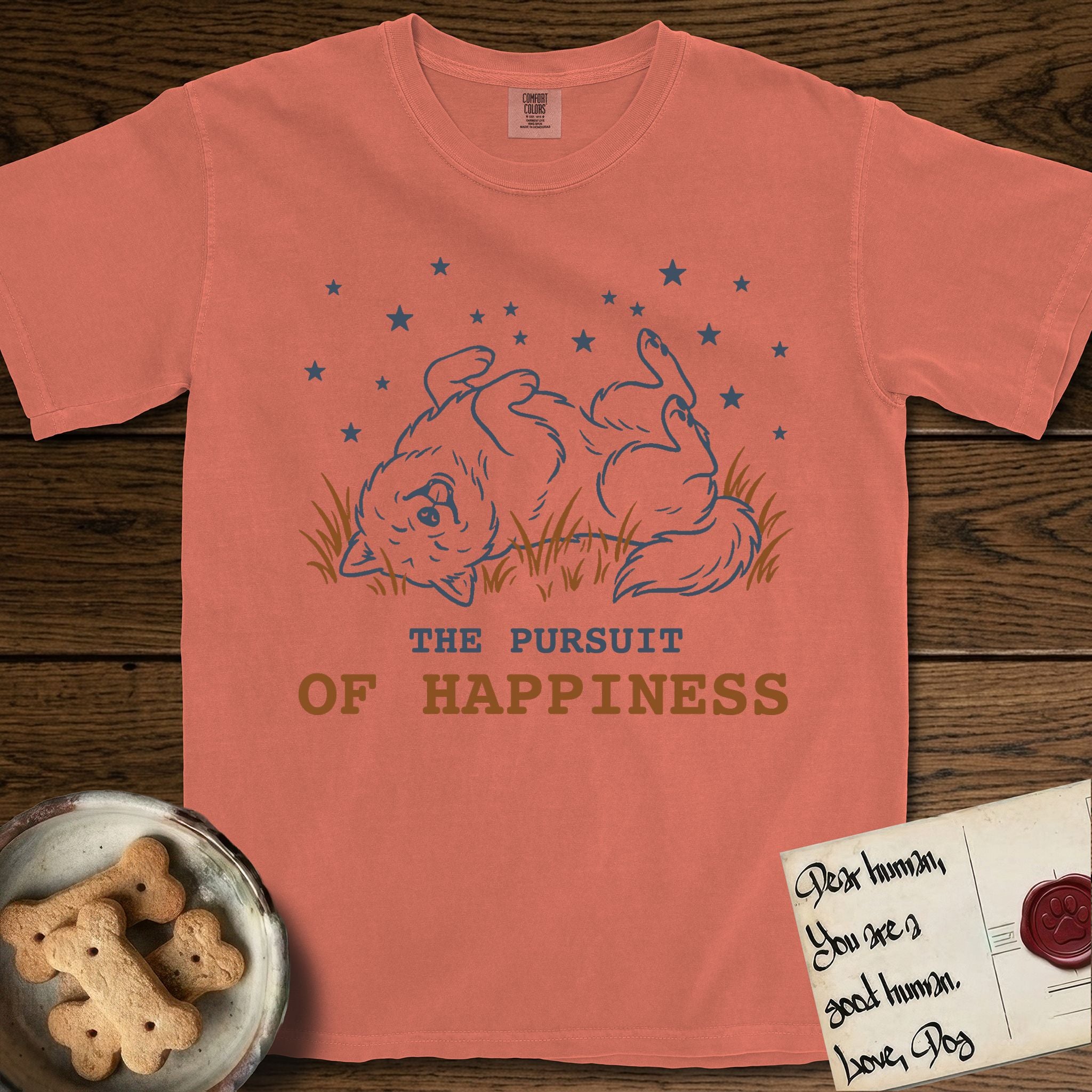 The Pursuit of Happiness T-Shirt flat lay in Terracotta, featuring a faded vintage-Americana line-art Samoyed rolling on his back in the grass with paws in the air under a sky of stars, above “The Pursuit of Happiness” lettering.