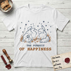 The Pursuit of Happiness T-Shirt flat lay in White, featuring a faded vintage-Americana line-art Samoyed rolling on his back in the grass with paws in the air under a sky of stars, above “The Pursuit of Happiness” lettering.