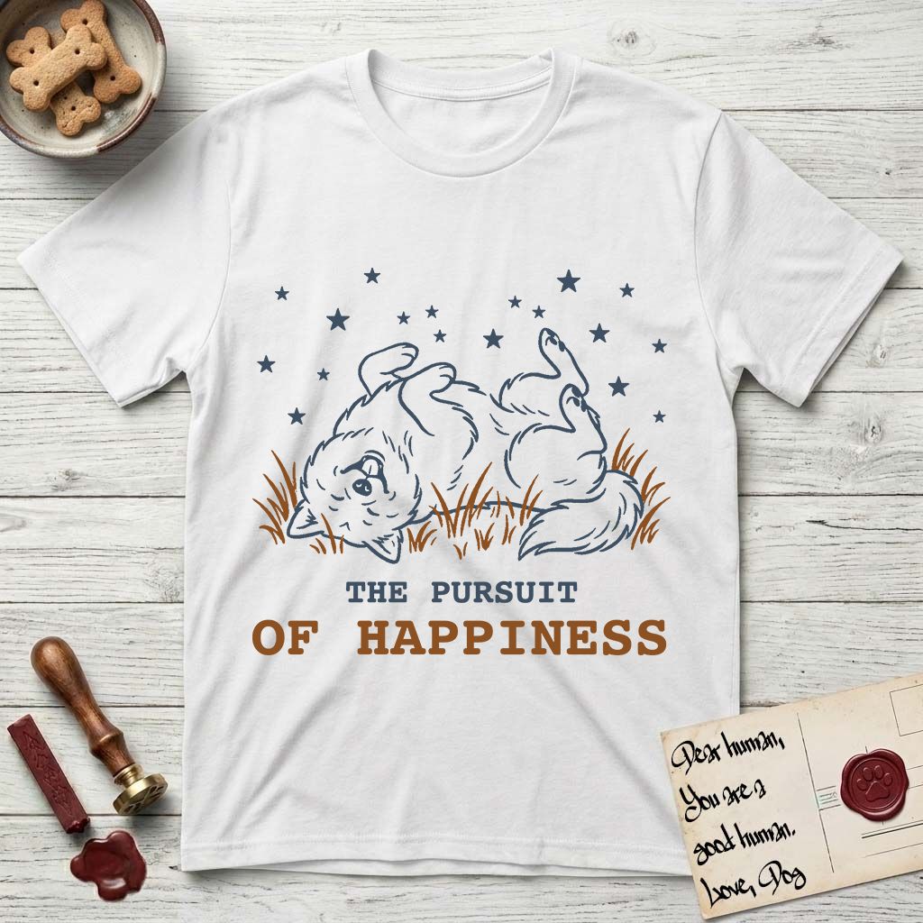 The Pursuit of Happiness T-Shirt flat lay in White, featuring a faded vintage-Americana line-art Samoyed rolling on his back in the grass with paws in the air under a sky of stars, above “The Pursuit of Happiness” lettering.