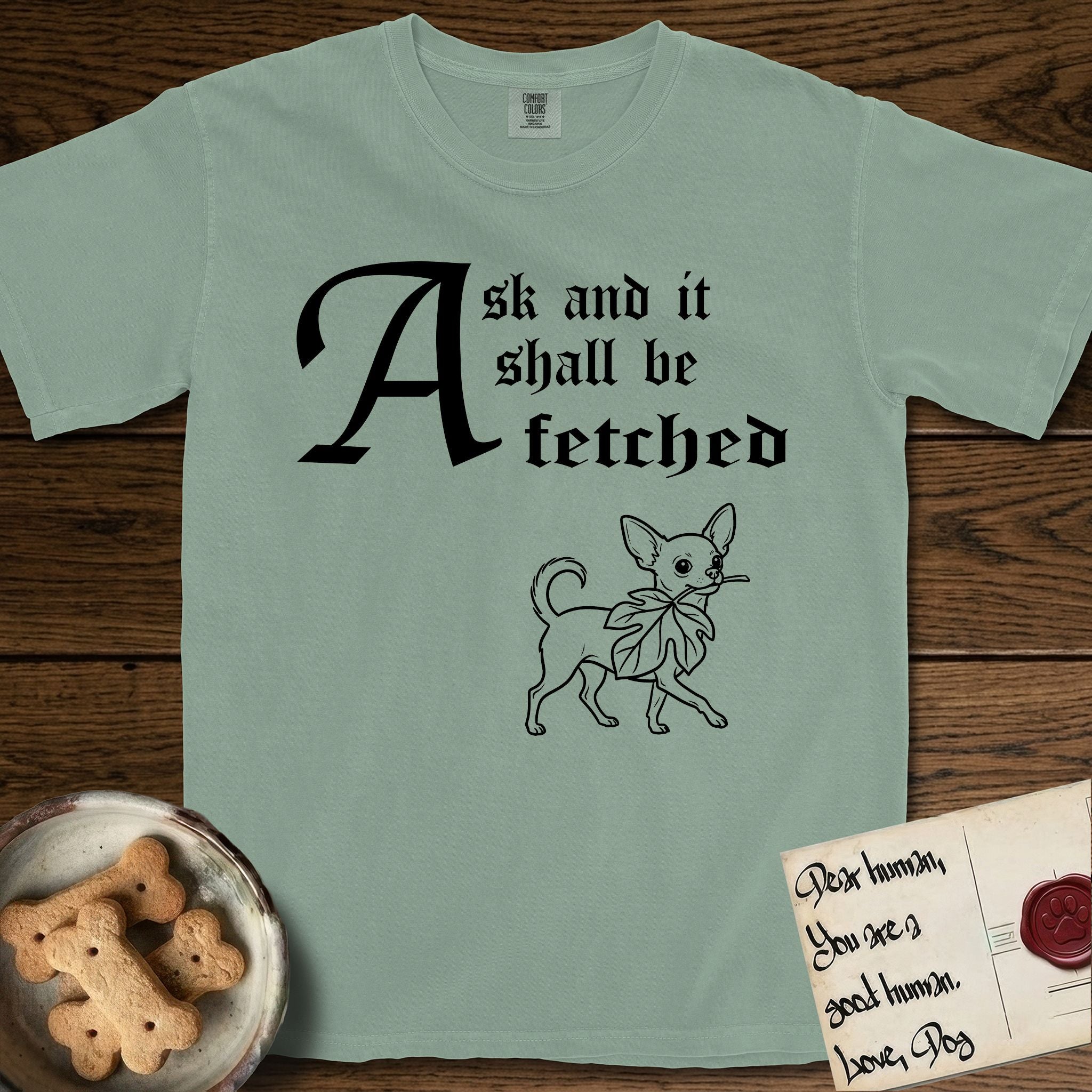 Ask and It Shall Be Fetched Chihuahua T-Shirt flat lay in Bay, featuring a sparse pen-and-ink illustration of a Chihuahua at a proud trot carrying an oversized fig leaf.