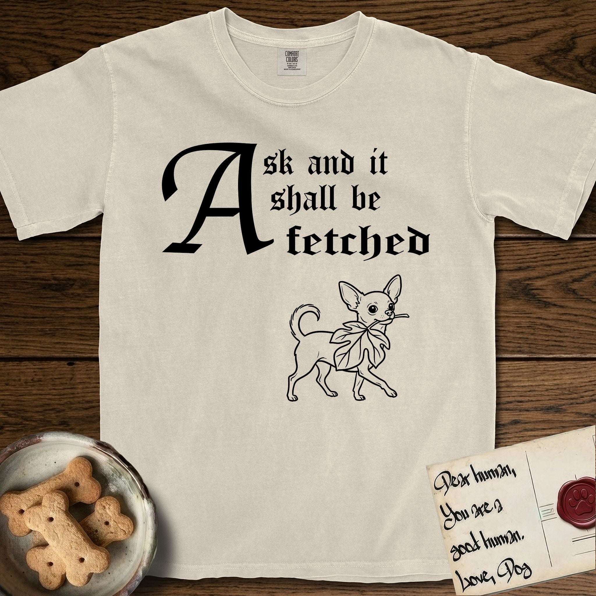 Ask and It Shall Be Fetched Chihuahua T-Shirt flat lay in Ivory, featuring a sparse pen-and-ink illustration of a Chihuahua at a proud trot carrying an oversized fig leaf.