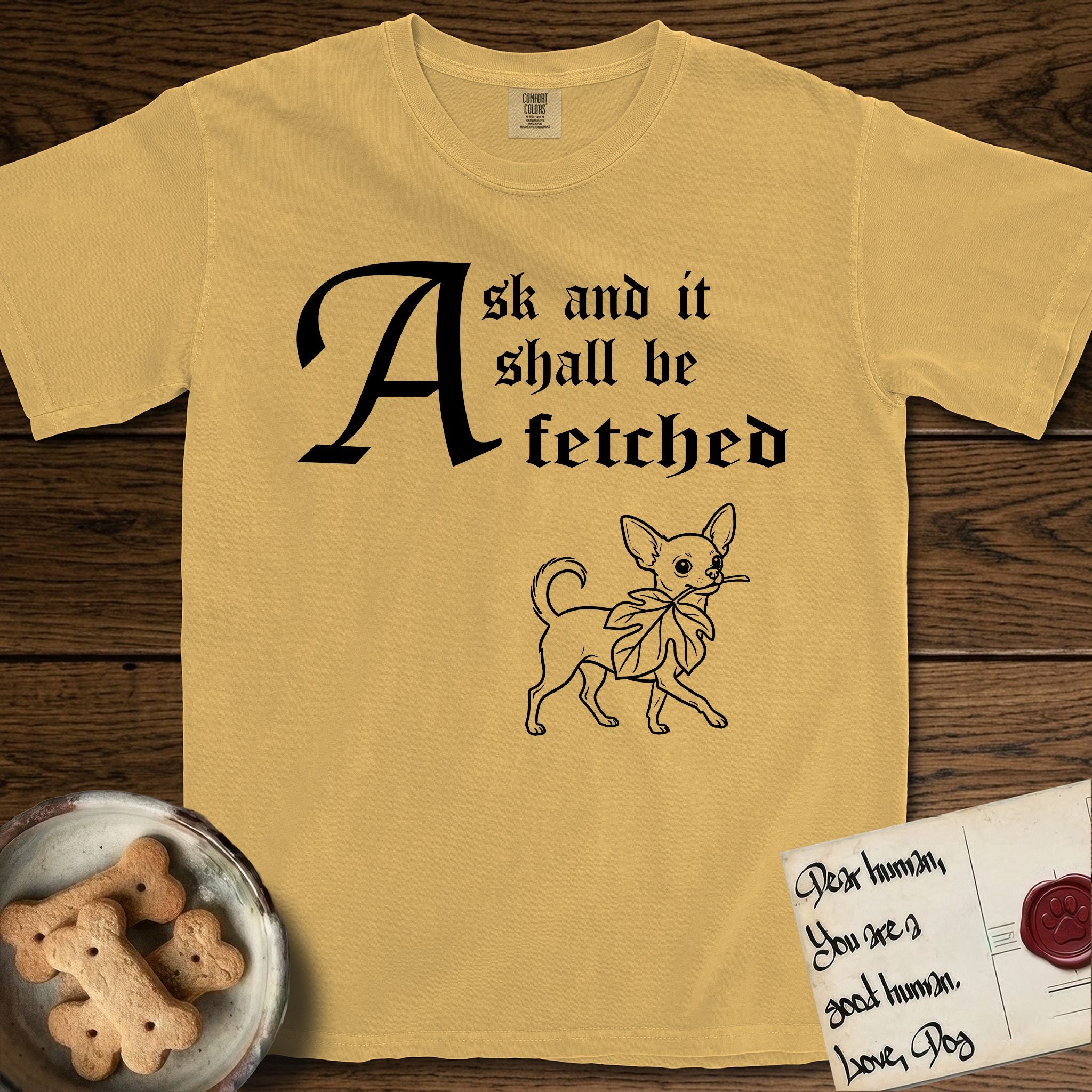 Ask and It Shall Be Fetched Chihuahua T-Shirt flat lay in Mustard, featuring a sparse pen-and-ink illustration of a Chihuahua at a proud trot carrying an oversized fig leaf.