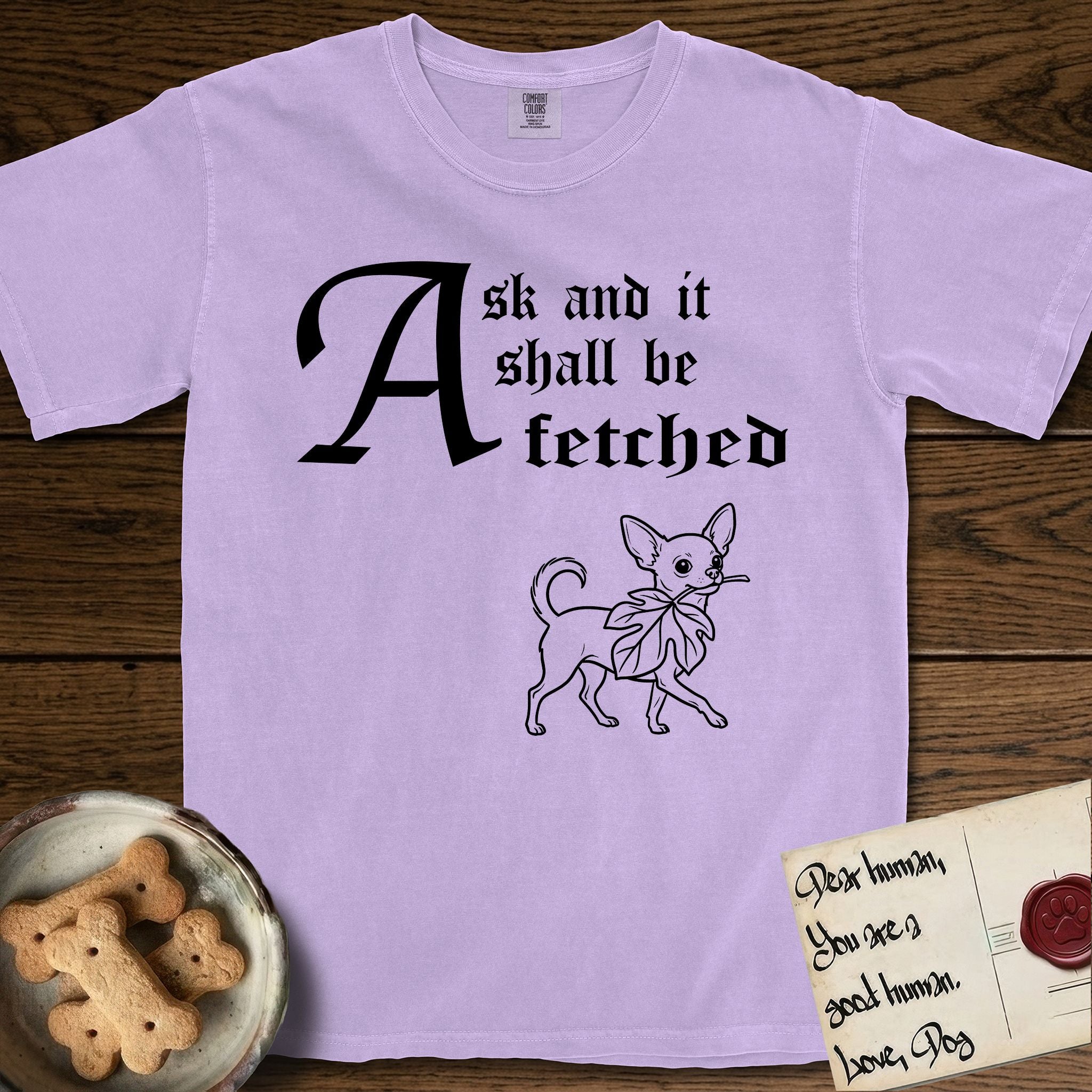 Ask and It Shall Be Fetched Chihuahua T-Shirt flat lay in Orchid, featuring a sparse pen-and-ink illustration of a Chihuahua at a proud trot carrying an oversized fig leaf.