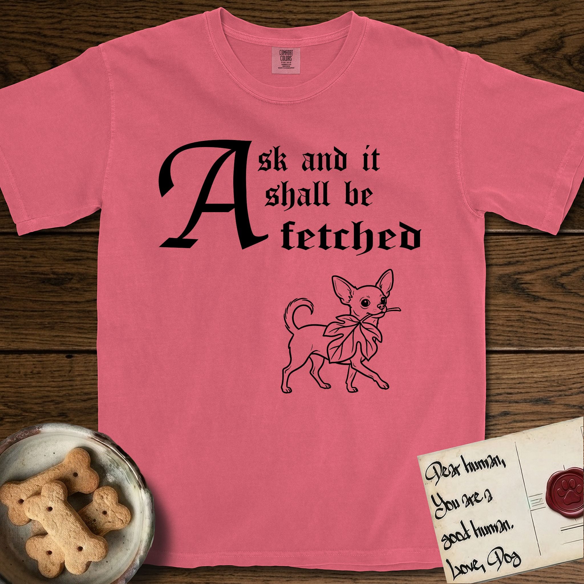 Ask and It Shall Be Fetched Chihuahua T-Shirt flat lay in Watermelon, featuring a sparse pen-and-ink illustration of a Chihuahua at a proud trot carrying an oversized fig leaf.