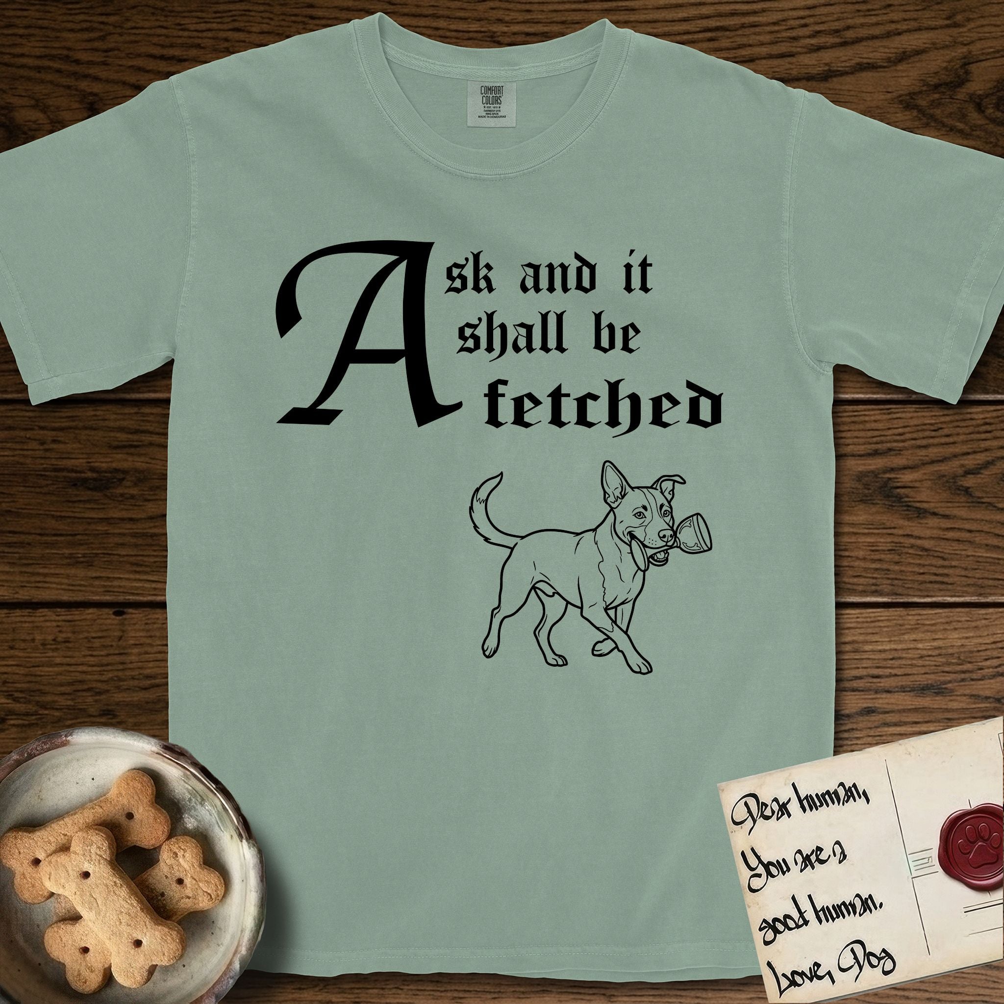 Ask and It Shall Be Fetched Lobo T-Shirt flat lay in Bay, featuring a sparse pen-and-ink illustration of Lobo, a black-and-white Australian Cattle Dog and Pitbull mix, at a proud trot carrying a chalice.