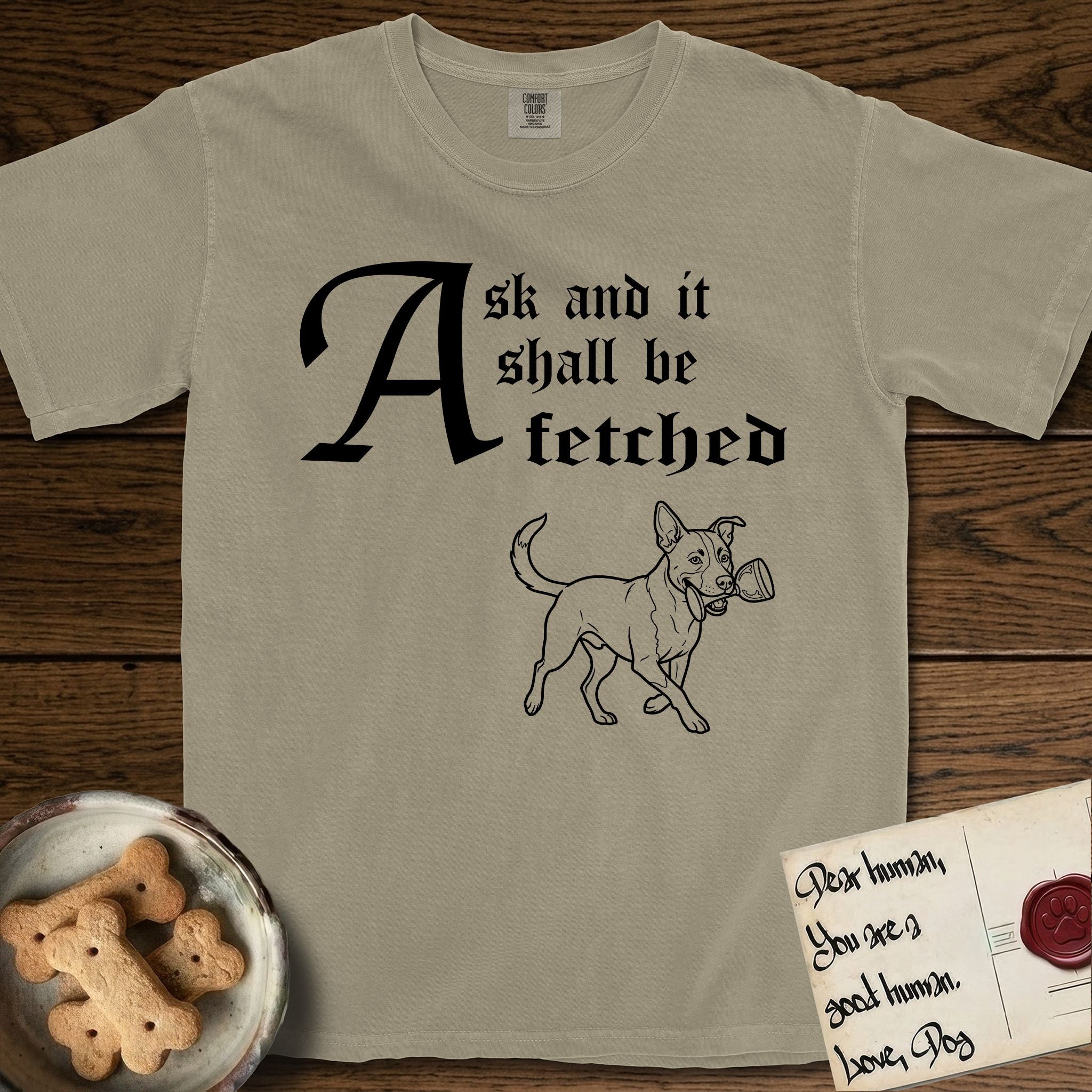 Ask and It Shall Be Fetched Lobo T-Shirt flat lay in Khaki, featuring a sparse pen-and-ink illustration of Lobo, a black-and-white Australian Cattle Dog and Pitbull mix, at a proud trot carrying a chalice.