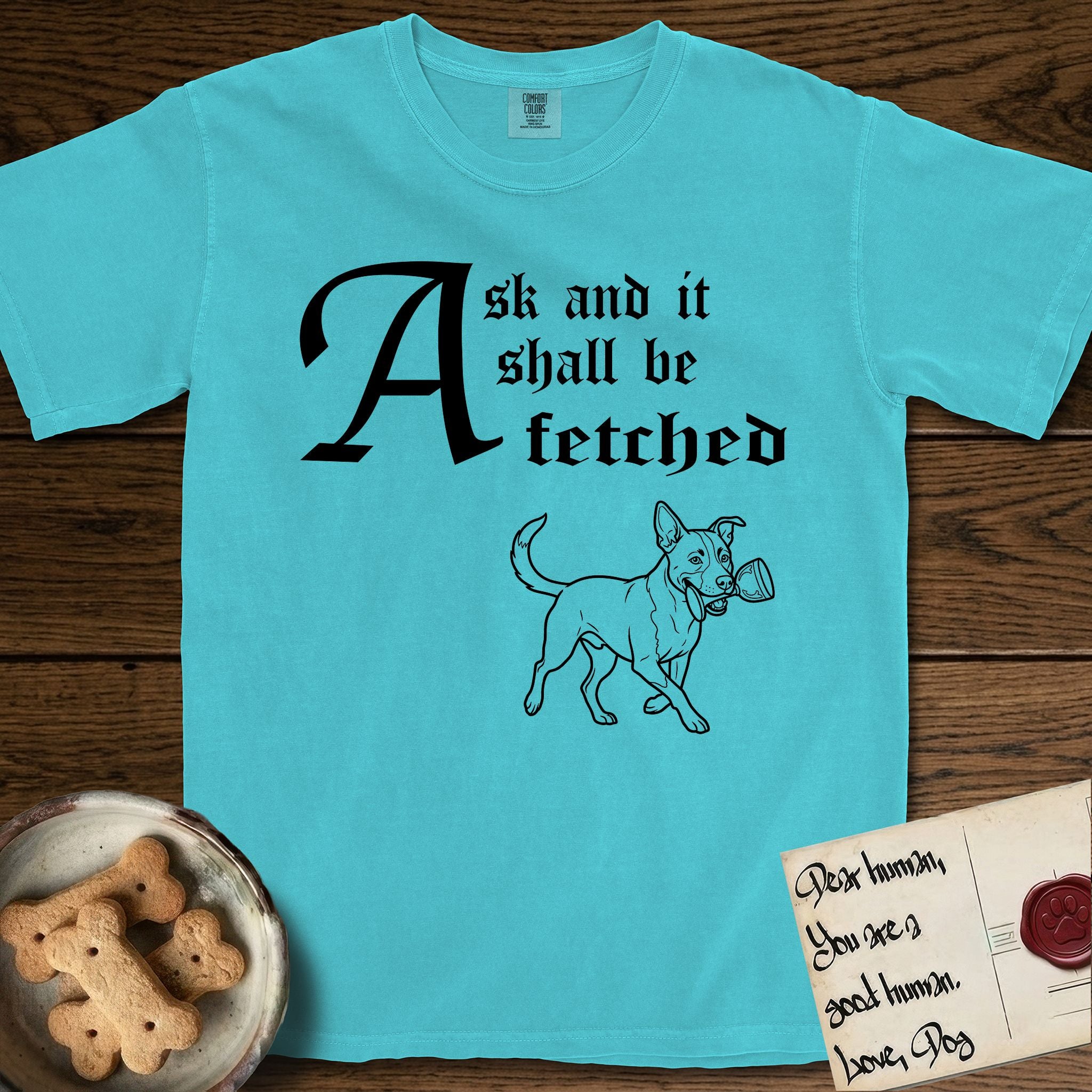 Ask and It Shall Be Fetched Lobo T-Shirt flat lay in Lagoon Blue, featuring a sparse pen-and-ink illustration of Lobo, a black-and-white Australian Cattle Dog and Pitbull mix, at a proud trot carrying a chalice.