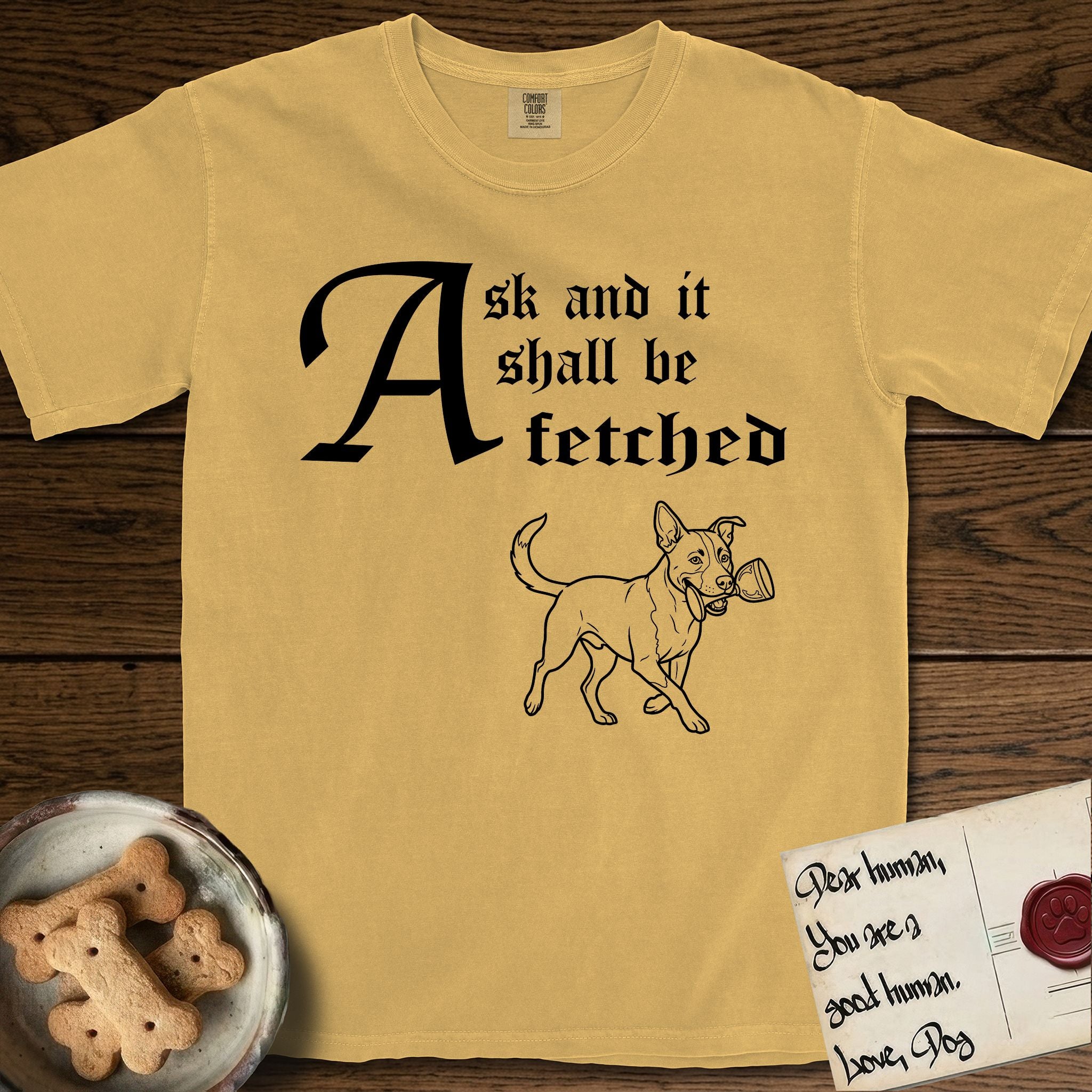 Ask and It Shall Be Fetched Lobo T-Shirt flat lay in Mustard, featuring a sparse pen-and-ink illustration of Lobo, a black-and-white Australian Cattle Dog and Pitbull mix, at a proud trot carrying a chalice.