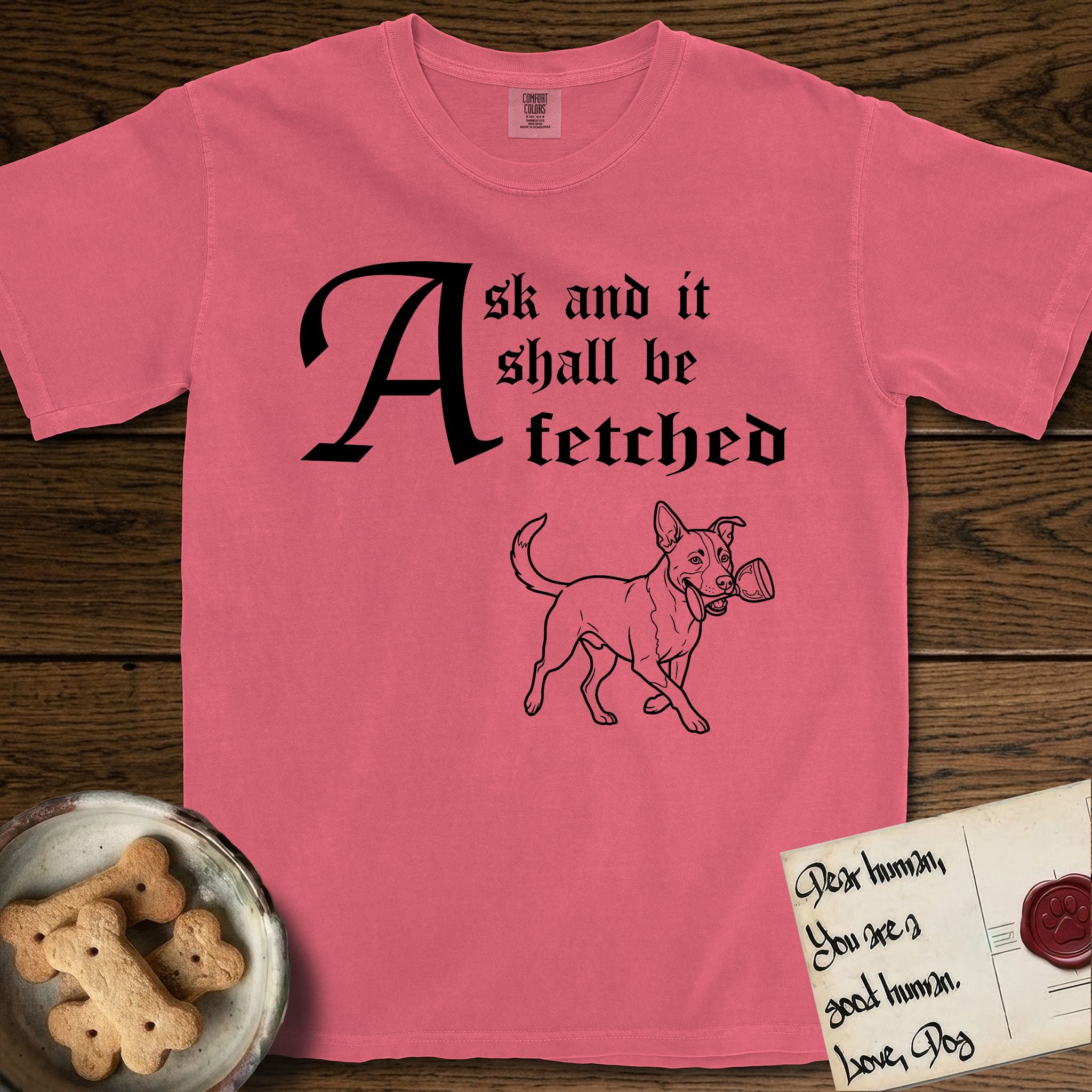 Ask and It Shall Be Fetched Lobo T-Shirt flat lay in Watermelon, featuring a sparse pen-and-ink illustration of Lobo, a black-and-white Australian Cattle Dog and Pitbull mix, at a proud trot carrying a chalice.
