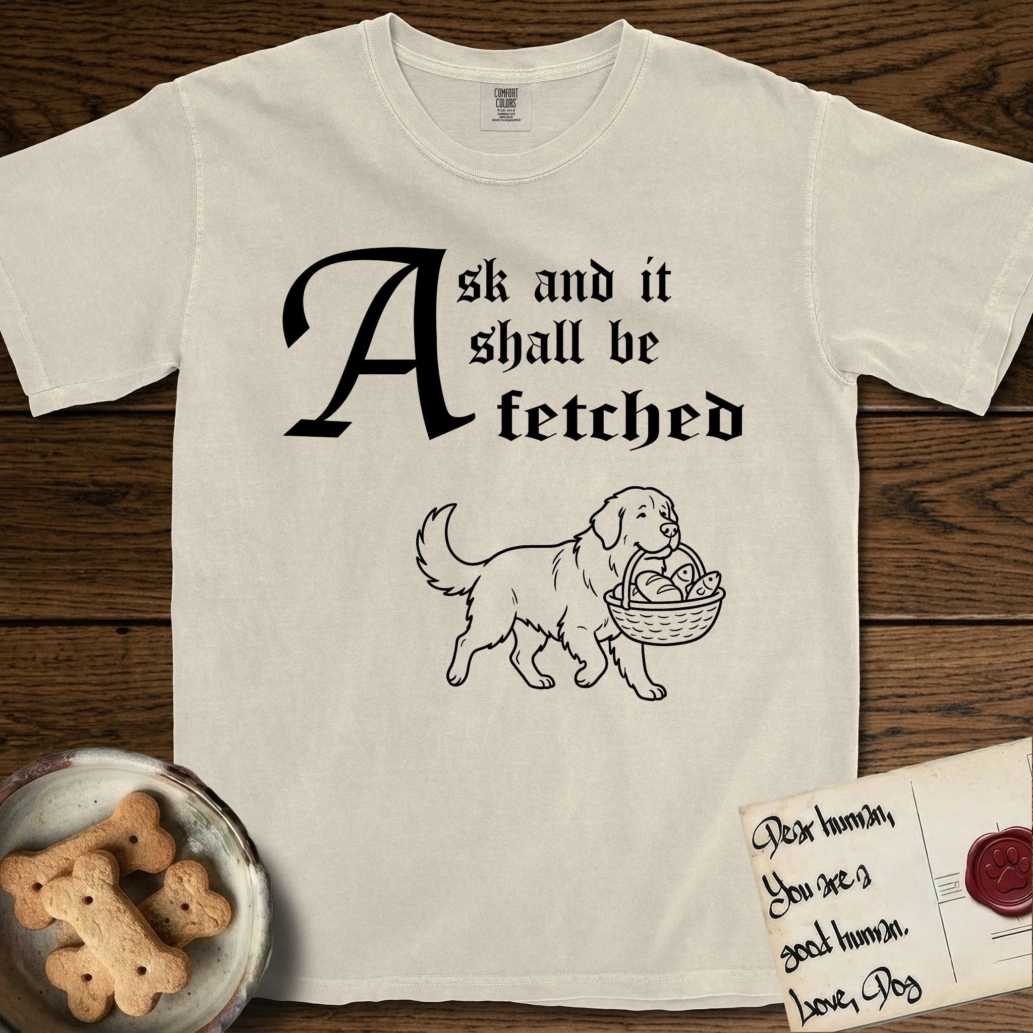 Ask and It Shall Be Fetched Newfoundland T-Shirt flat lay in Ivory, featuring a sparse pen-and-ink illustration of a Newfoundland at a proud trot carrying loaves and fishes.