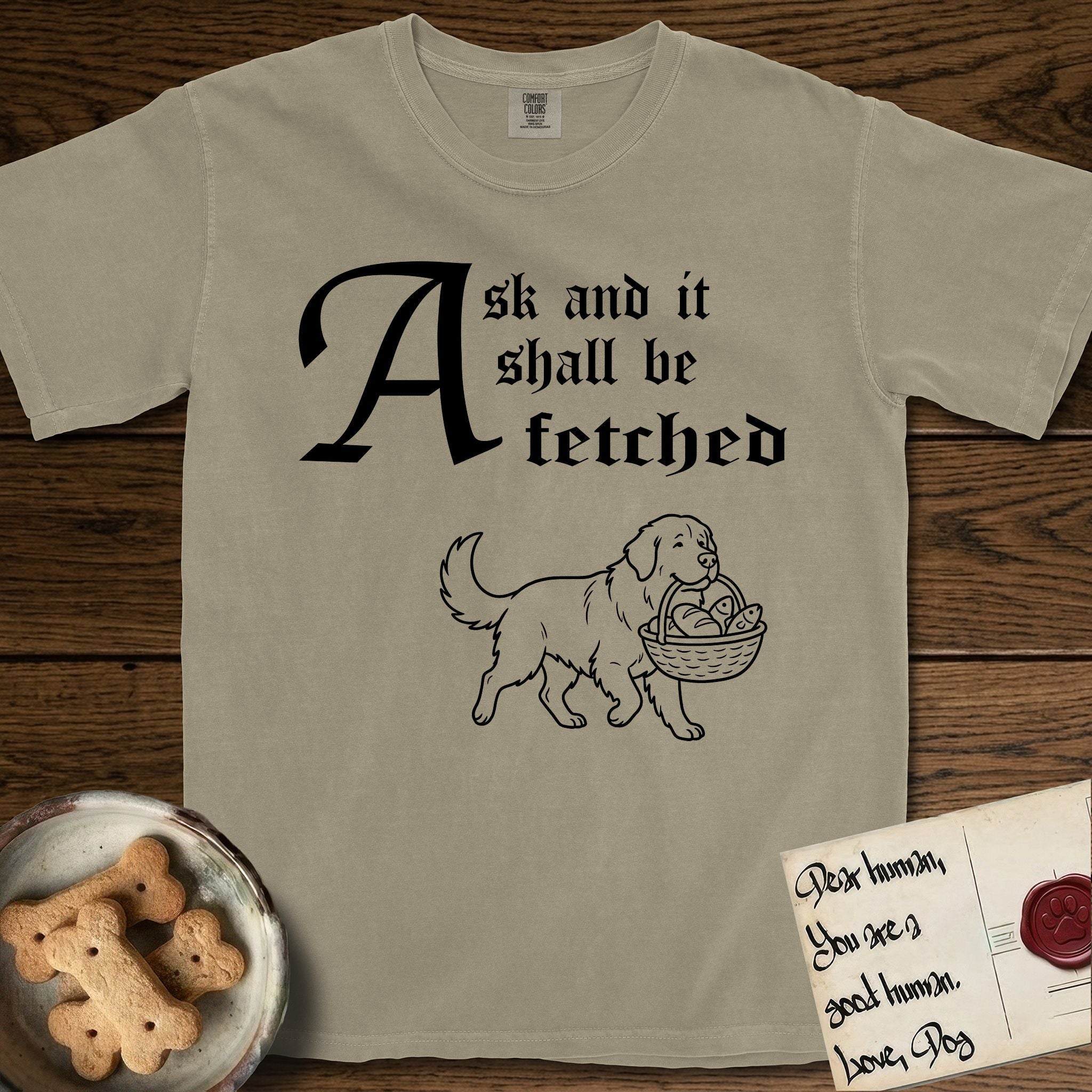 Ask and It Shall Be Fetched Newfoundland T-Shirt flat lay in Khaki, featuring a sparse pen-and-ink illustration of a Newfoundland at a proud trot carrying loaves and fishes.