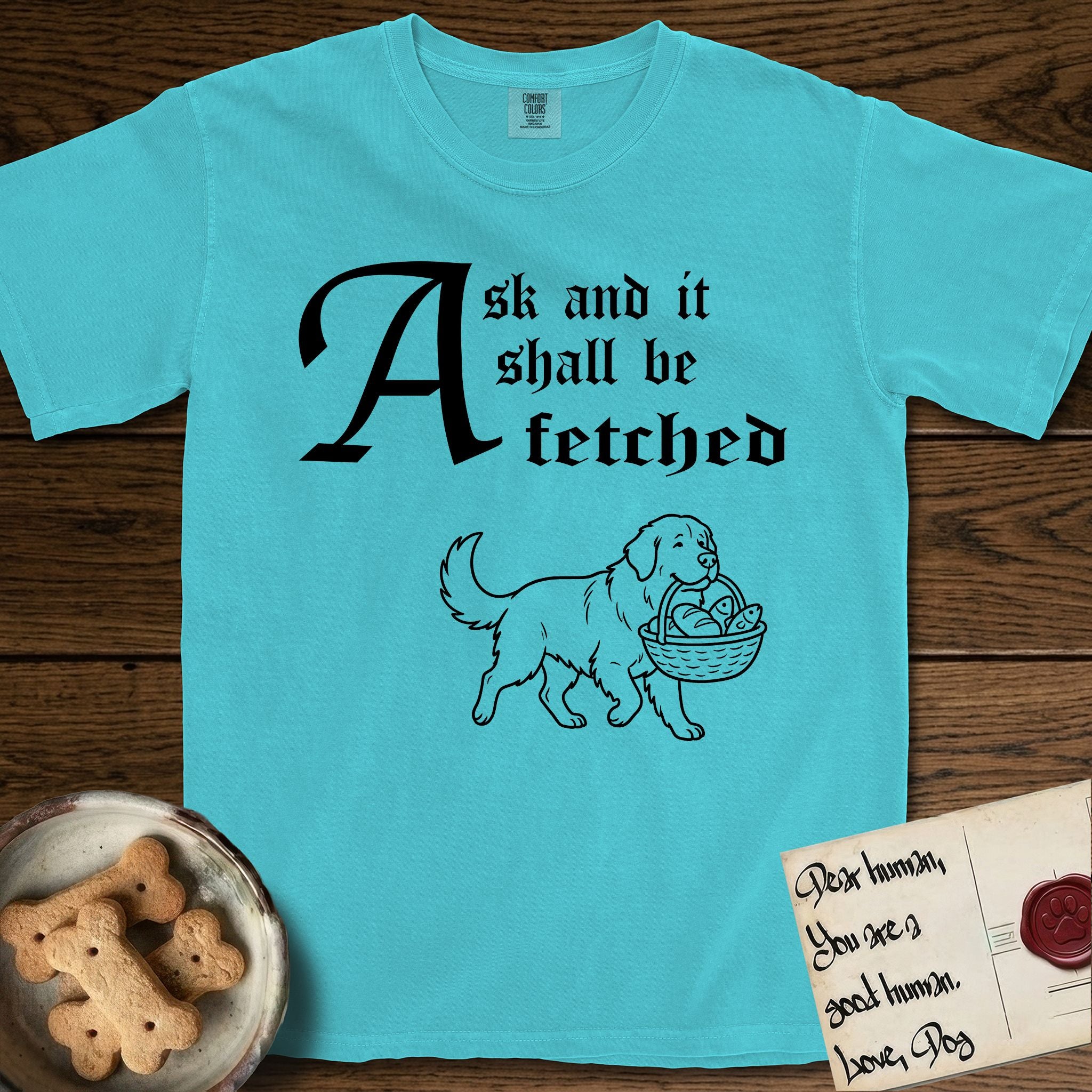 Ask and It Shall Be Fetched Newfoundland T-Shirt flat lay in Lagoon Blue, featuring a sparse pen-and-ink illustration of a Newfoundland at a proud trot carrying loaves and fishes.