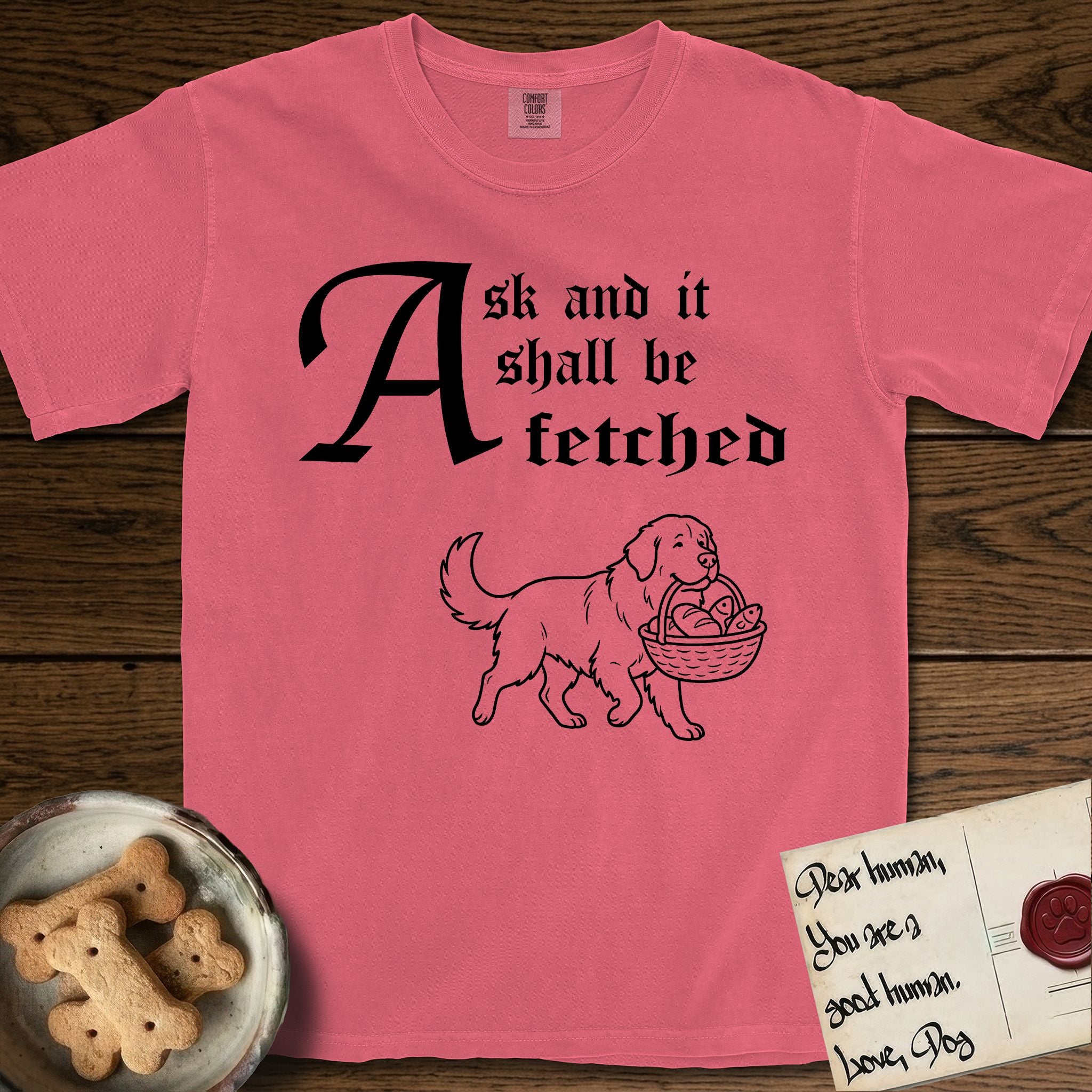 Ask and It Shall Be Fetched Newfoundland T-Shirt flat lay in Watermelon, featuring a sparse pen-and-ink illustration of a Newfoundland at a proud trot carrying loaves and fishes.