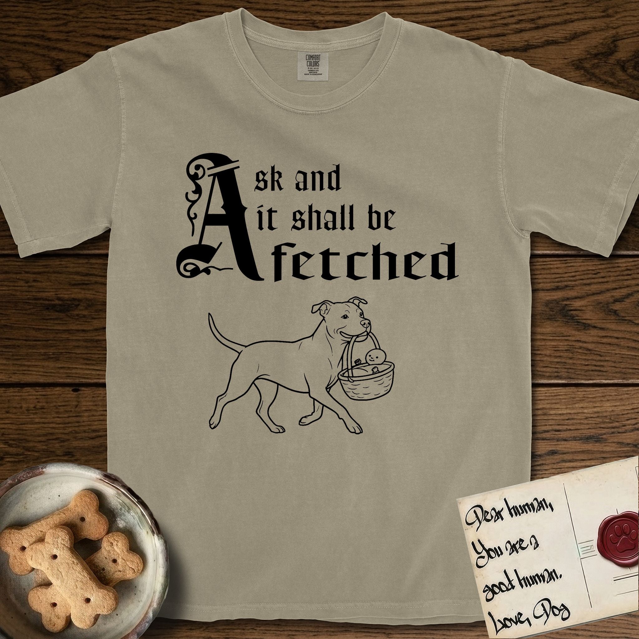 Ask and It Shall Be Fetched Pitbull T-Shirt flat lay in Khaki, featuring a sparse pen-and-ink illustration of a Pitbull at a proud trot carrying a baby in a reed basket.