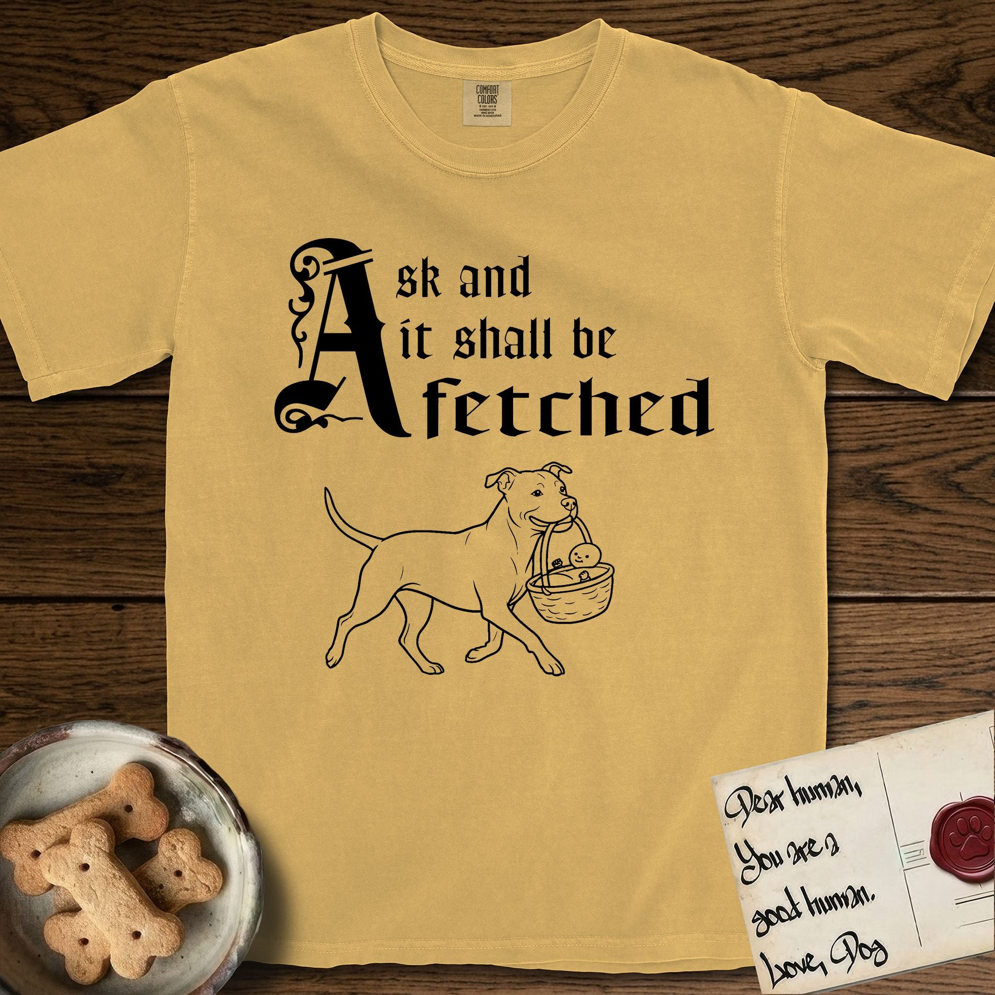 Ask and It Shall Be Fetched Pitbull T-Shirt flat lay in Mustard, featuring a sparse pen-and-ink illustration of a Pitbull at a proud trot carrying a baby in a reed basket.