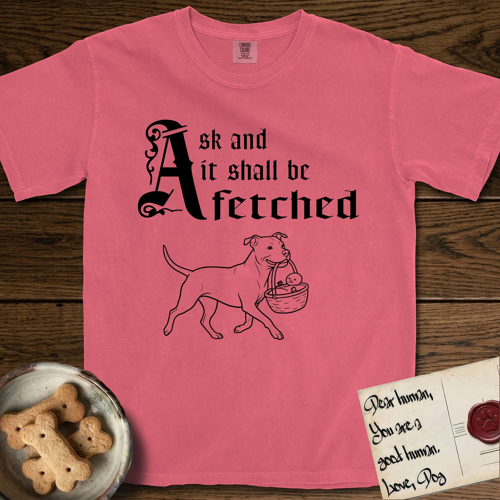 Ask and It Shall Be Fetched Pitbull T-Shirt flat lay in Watermelon, featuring a sparse pen-and-ink illustration of a Pitbull at a proud trot carrying a baby in a reed basket.