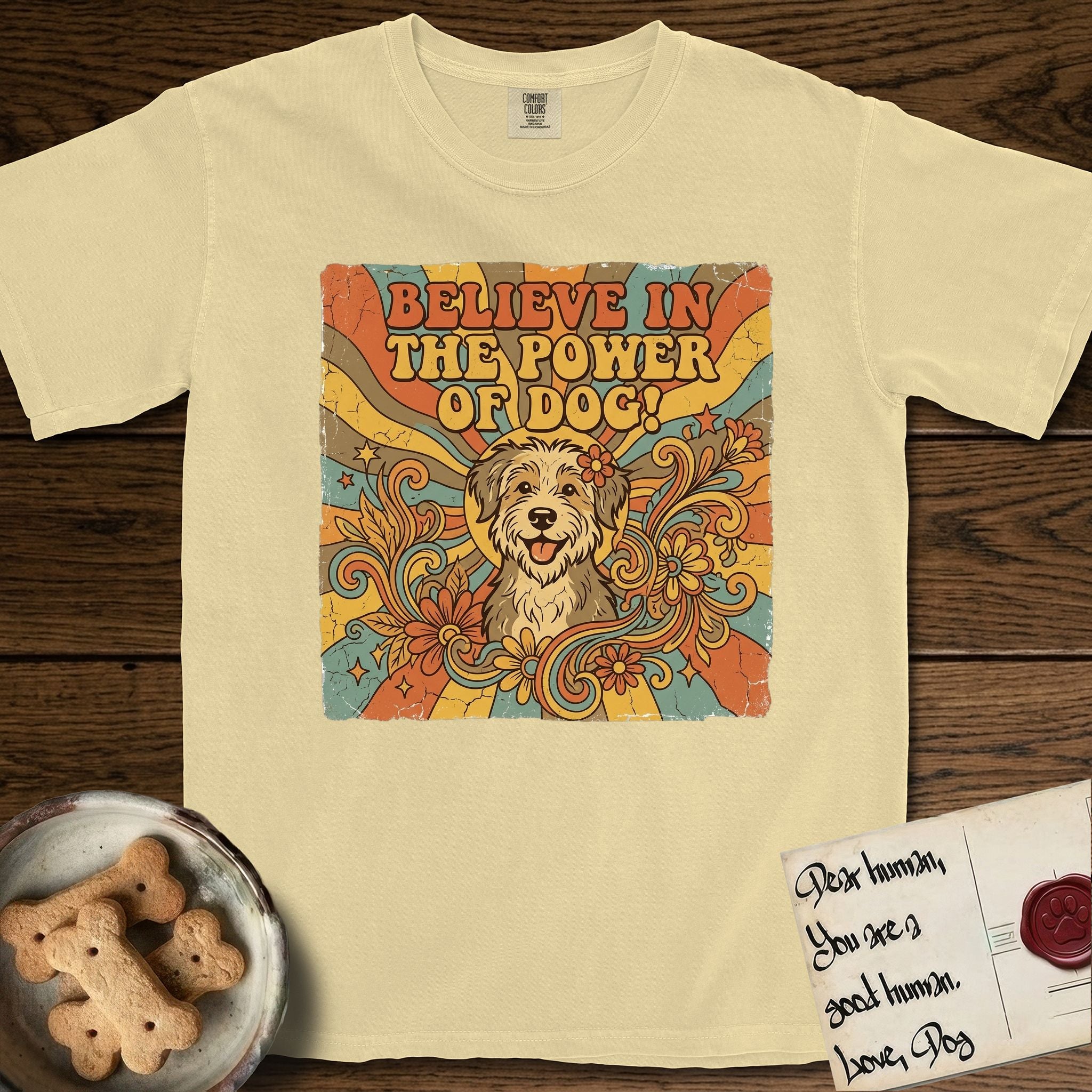 Believe in the Power of Dog Rescue Mutt T-Shirt flat lay in Butter, featuring a retro 1970s groovy poster of a grinning rescue mutt with a radiating sunburst and swirling florals.