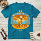 Believe in the Power of Dog Yellow Lab T-Shirt flat lay in Tropical Blue, featuring a vintage oval badge with a haloed Yellow Lab running through a field of wildflowers and the words Joy, Loyalty, Love.
