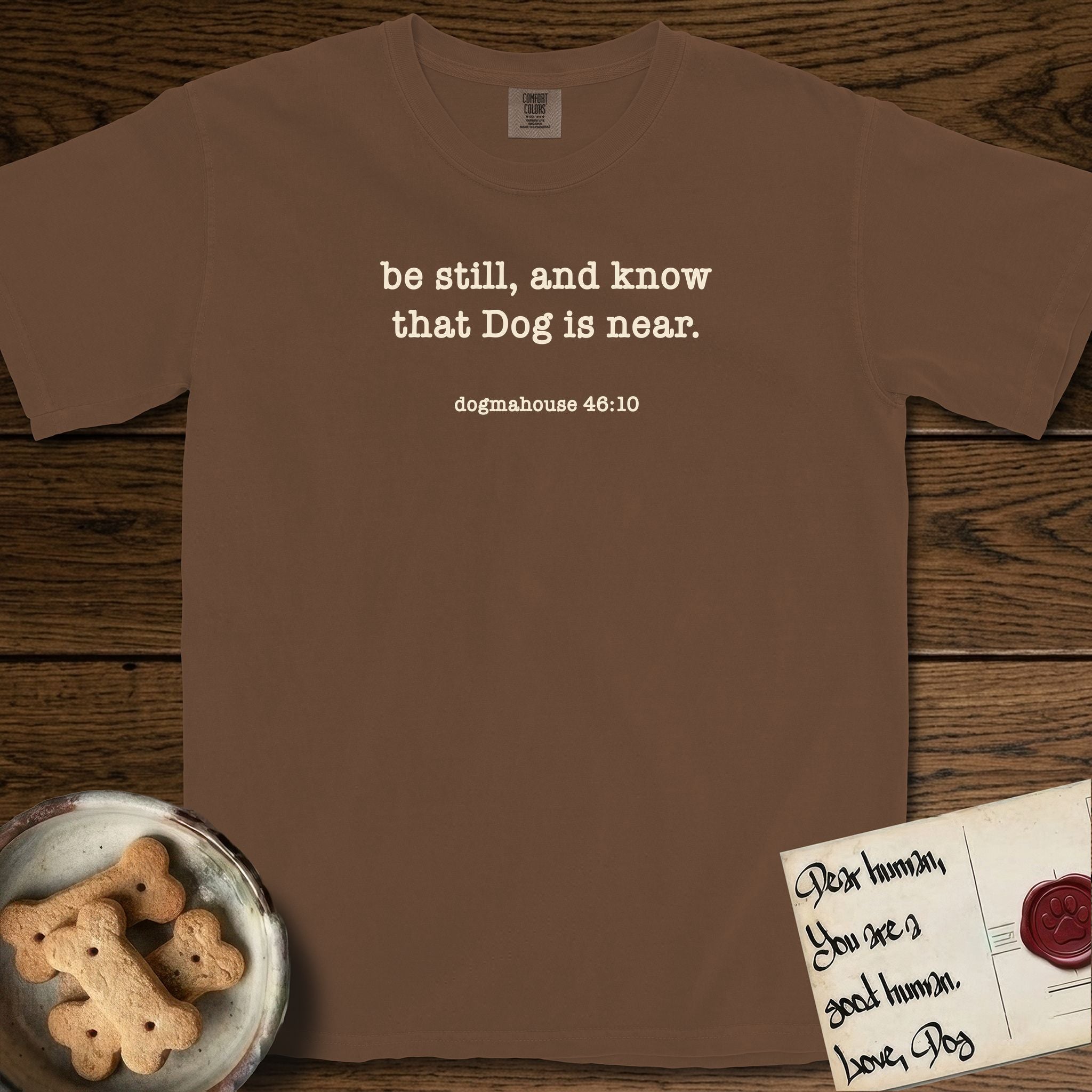 Be Still T-Shirt flat lay in Espresso, featuring Small centered typewriter-type text reading be still, and know that Dog is near, with the citation dogmahouse 46:10 beneath it.