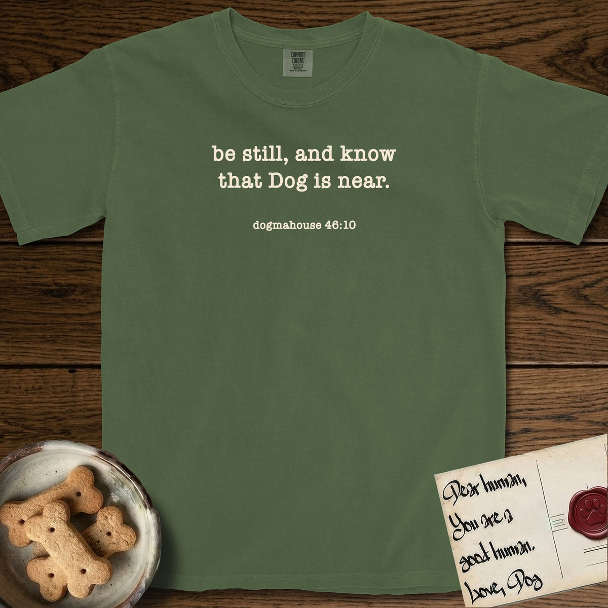 Be Still T-Shirt flat lay in Moss, featuring Small centered typewriter-type text reading be still, and know that Dog is near, with the citation dogmahouse 46:10 beneath it.