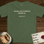 Knock T-Shirt flat lay in Moss, featuring Small centered typewriter-type text reading knock, and it shall be barked at, with the citation dogmahouse 7:7 beneath it.