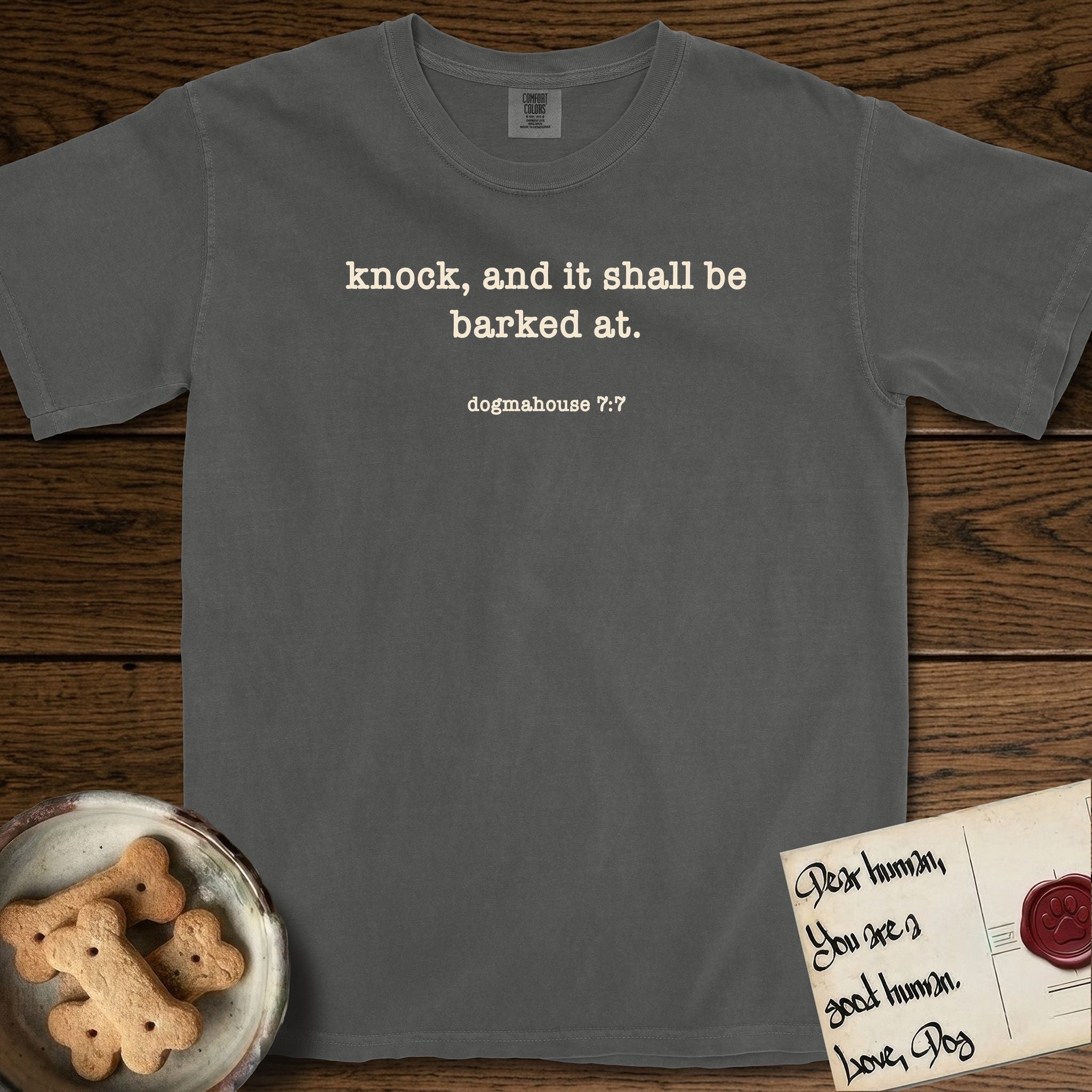 Knock T-Shirt flat lay in Pepper, featuring Small centered typewriter-type text reading knock, and it shall be barked at, with the citation dogmahouse 7:7 beneath it.