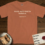 Knock T-Shirt flat lay in Yam, featuring Small centered typewriter-type text reading knock, and it shall be barked at, with the citation dogmahouse 7:7 beneath it.