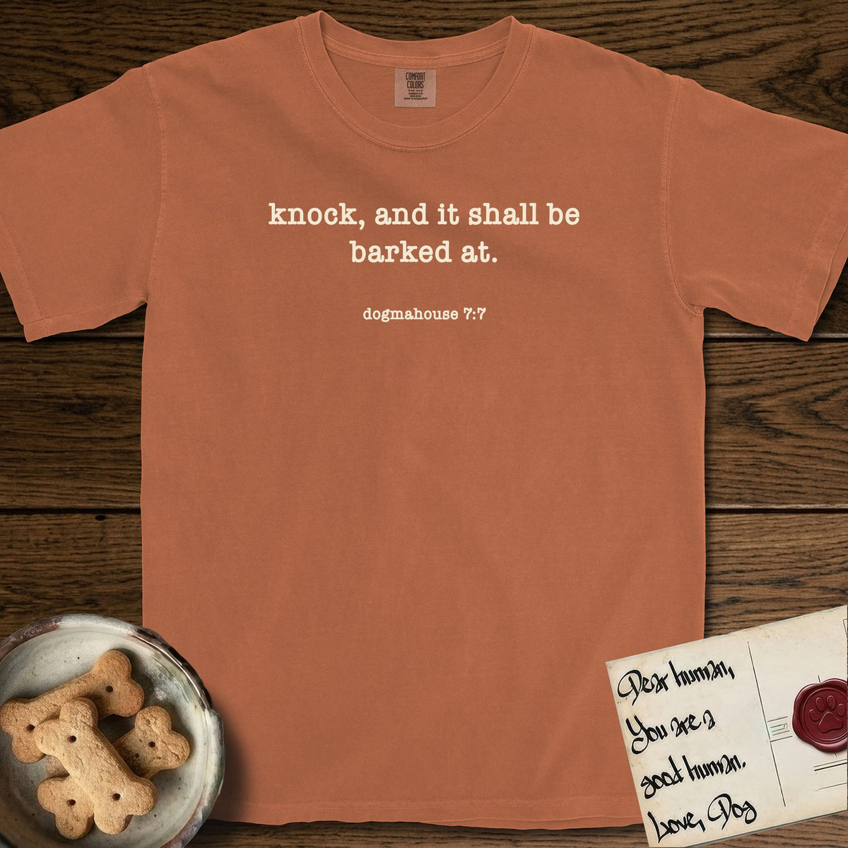 Knock T-Shirt flat lay in Yam, featuring Small centered typewriter-type text reading knock, and it shall be barked at, with the citation dogmahouse 7:7 beneath it.