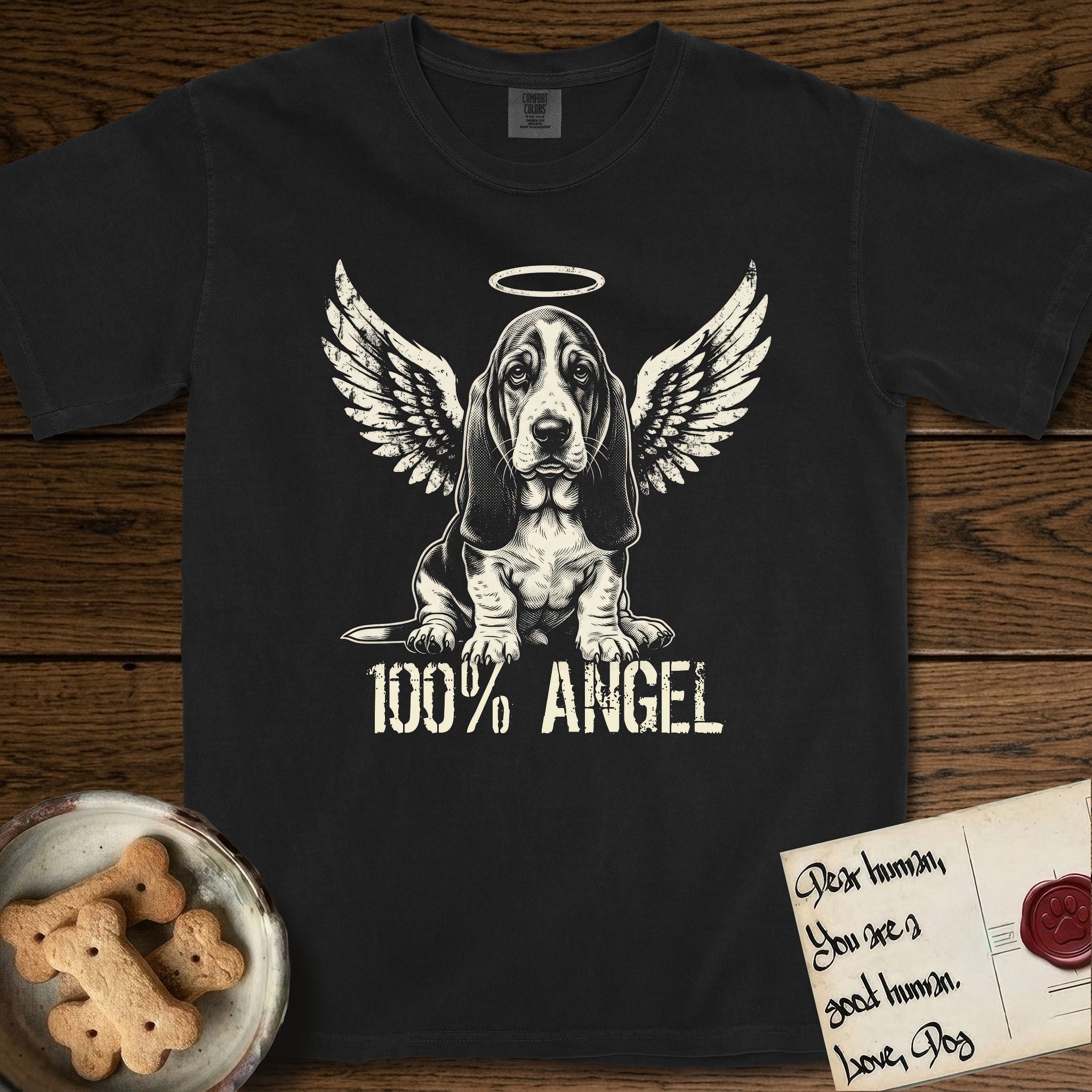 Basset Hound T-Shirt flat lay in Black, featuring Distressed cream one-color screen-print of a Basset Hound puppy as an angel, long ears draping forward, wings spread and a halo above, with 100% ANGEL lettering across the bottom.