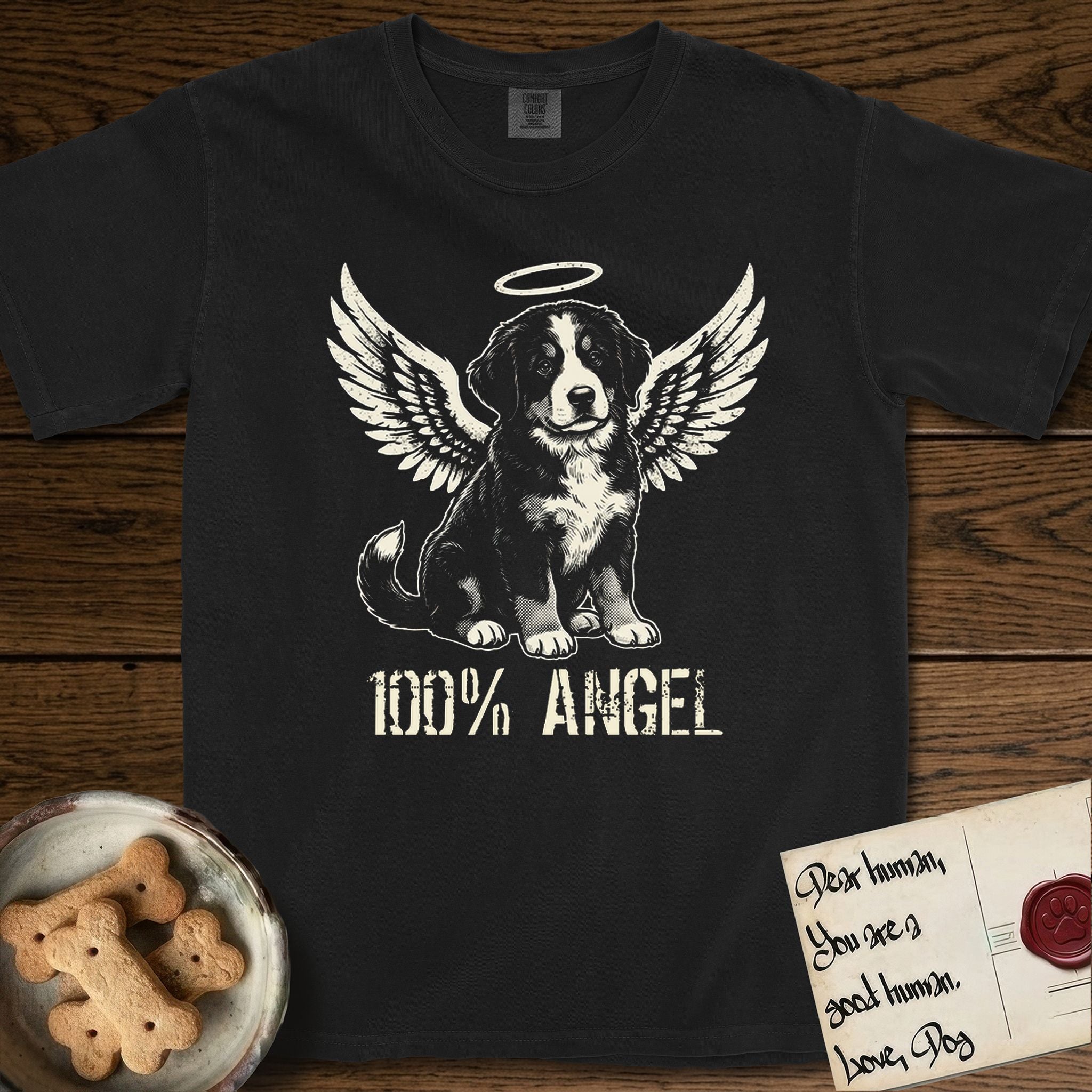 Bernese Mountain Dog T-Shirt flat lay in Black, featuring Distressed cream one-color screen-print of a Bernese Mountain Dog puppy as an angel, three-quarter sitting pose with bushy tail curled, wings spread and a halo above, with 100% ANGEL lettering across the bottom.
