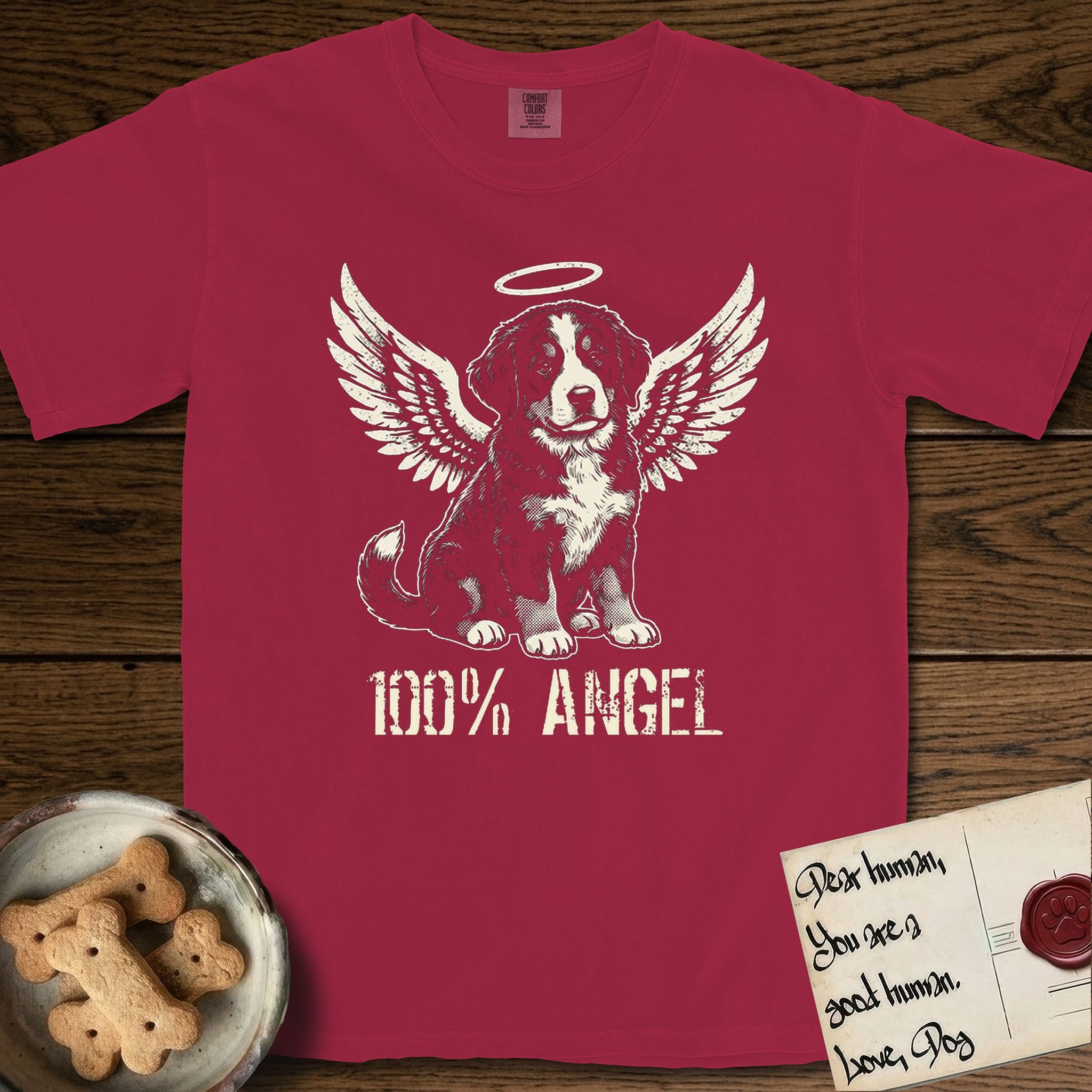 Bernese Mountain Dog T-Shirt flat lay in Chili, featuring Distressed cream one-color screen-print of a Bernese Mountain Dog puppy as an angel, three-quarter sitting pose with bushy tail curled, wings spread and a halo above, with 100% ANGEL lettering across the bottom.