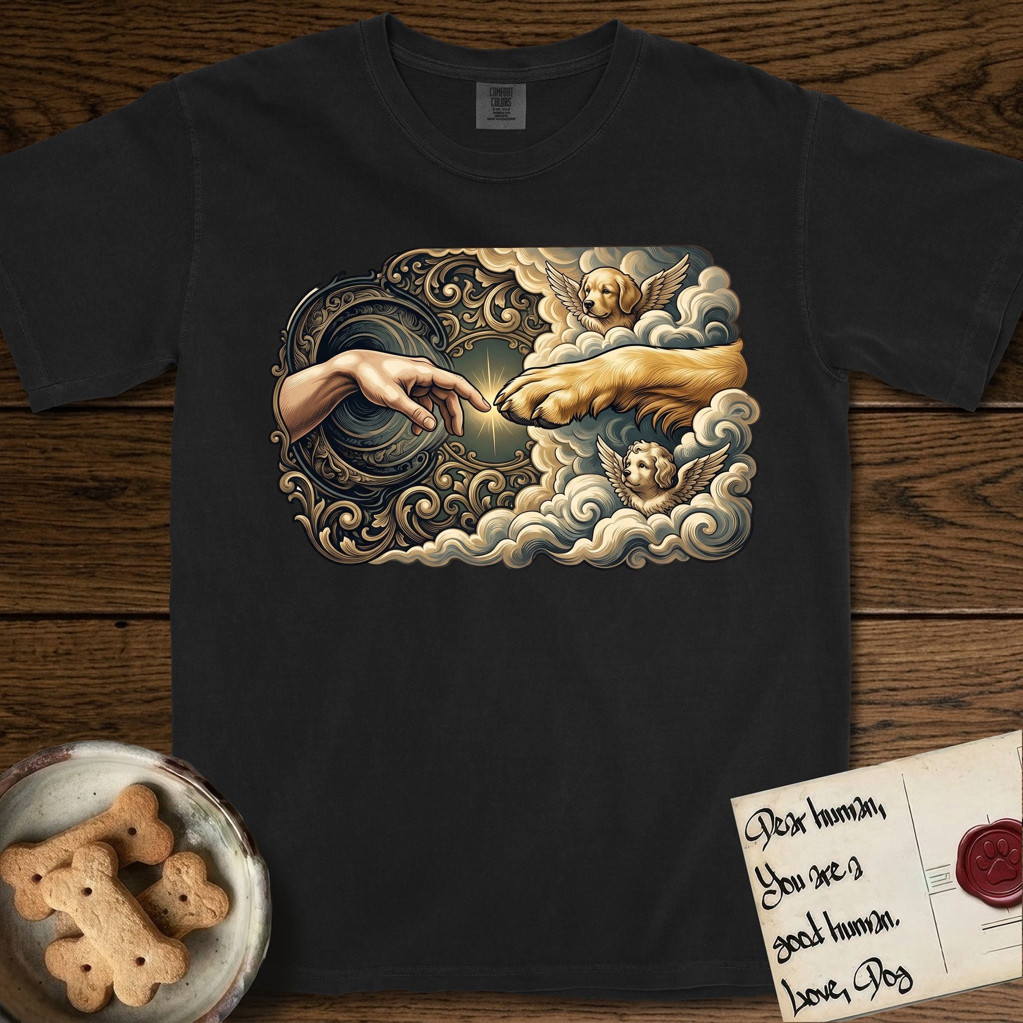Creation of Adam T-Shirt flat lay in Black, featuring a baroque ornamental frame with a Golden Retriever emerging from clouds extending a paw toward a reaching human hand, a small angel beside them, and a glowing point of contact.