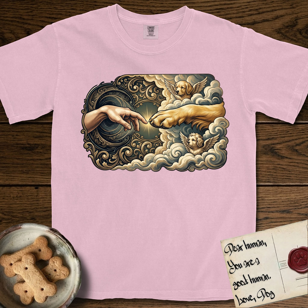 Creation of Adam T-Shirt flat lay in Blossom, featuring a baroque ornamental frame with a Golden Retriever emerging from clouds extending a paw toward a reaching human hand, a small angel beside them, and a glowing point of contact.