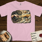 Creation of Adam T-Shirt flat lay in Blossom, featuring a baroque ornamental frame with a Golden Retriever emerging from clouds extending a paw toward a reaching human hand, a small angel beside them, and a glowing point of contact.