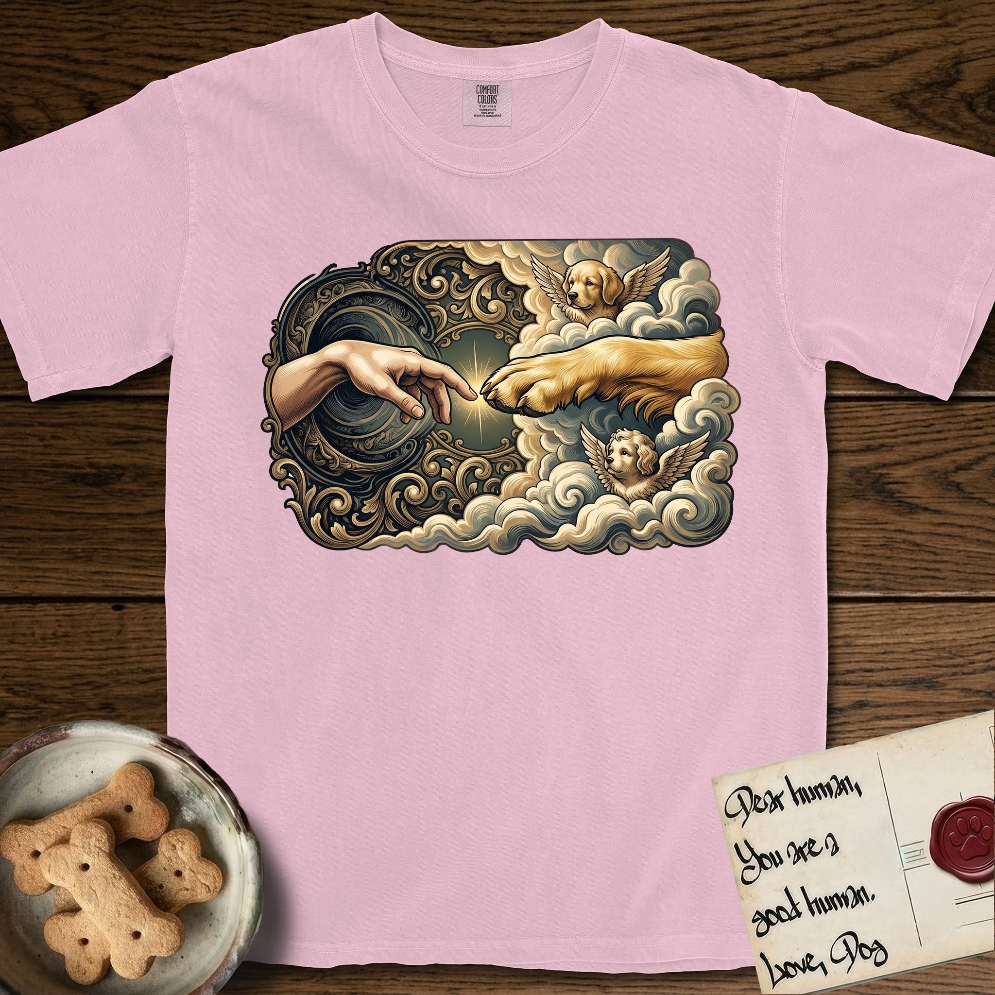 Creation of Adam T-Shirt flat lay in Blossom, featuring a baroque ornamental frame with a Golden Retriever emerging from clouds extending a paw toward a reaching human hand, a small angel beside them, and a glowing point of contact.