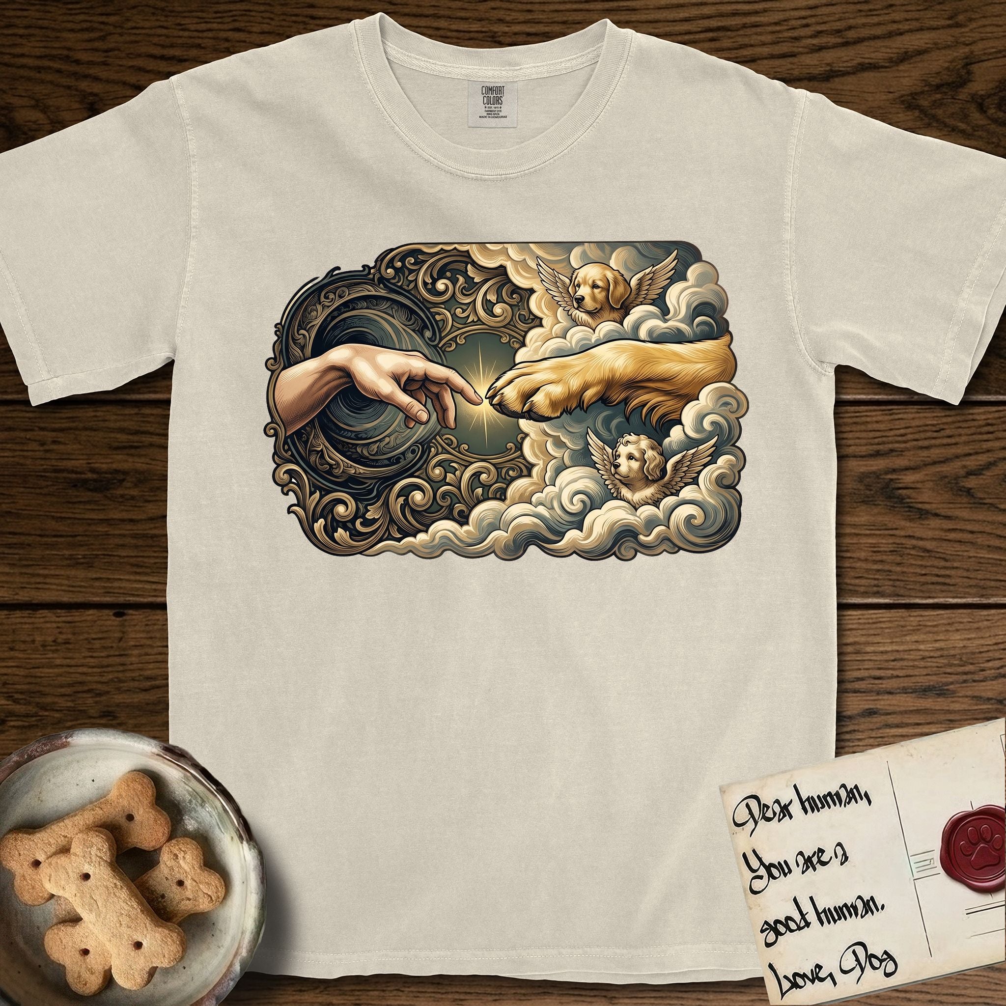 Creation of Adam T-Shirt flat lay in Ivory, featuring a baroque ornamental frame with a Golden Retriever emerging from clouds extending a paw toward a reaching human hand, a small angel beside them, and a glowing point of contact.