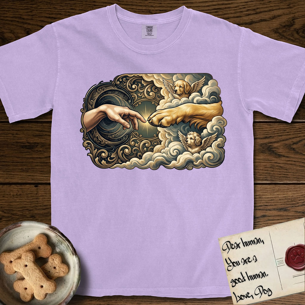 Creation of Adam T-Shirt flat lay in Orchid, featuring a baroque ornamental frame with a Golden Retriever emerging from clouds extending a paw toward a reaching human hand, a small angel beside them, and a glowing point of contact.