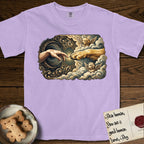 Creation of Adam T-Shirt flat lay in Orchid, featuring a baroque ornamental frame with a Golden Retriever emerging from clouds extending a paw toward a reaching human hand, a small angel beside them, and a glowing point of contact.