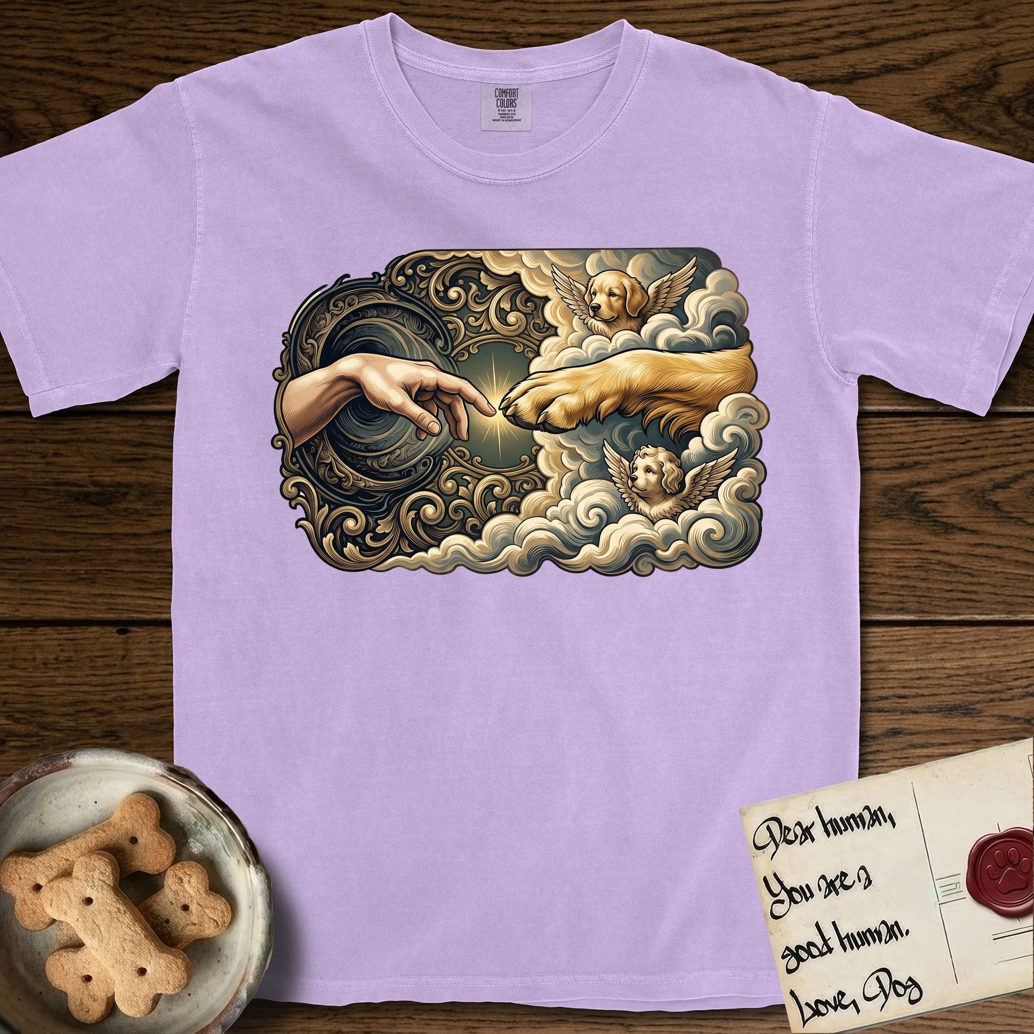 Creation of Adam T-Shirt flat lay in Orchid, featuring a baroque ornamental frame with a Golden Retriever emerging from clouds extending a paw toward a reaching human hand, a small angel beside them, and a glowing point of contact.