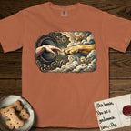 Creation of Adam T-Shirt flat lay in Yam, featuring a baroque ornamental frame with a Golden Retriever emerging from clouds extending a paw toward a reaching human hand, a small angel beside them, and a glowing point of contact.