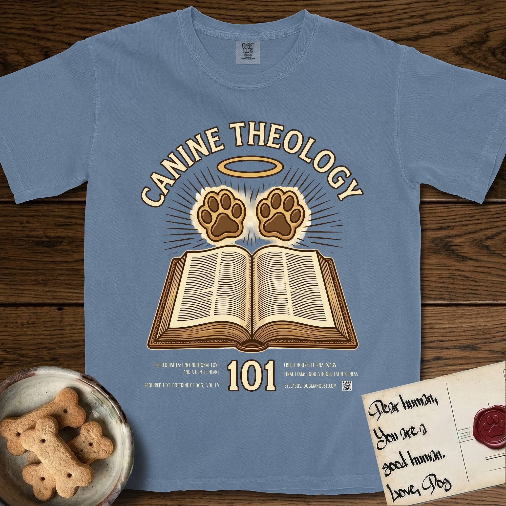 Canine Theology 101 T-Shirt flat lay in Blue Jean, featuring a cream badge with an open book and two glowing paw prints rising above it, ringed by a circular halo.