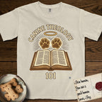 Canine Theology 101 T-Shirt flat lay in Ivory, featuring a cream badge with an open book and two glowing paw prints rising above it, ringed by a circular halo.