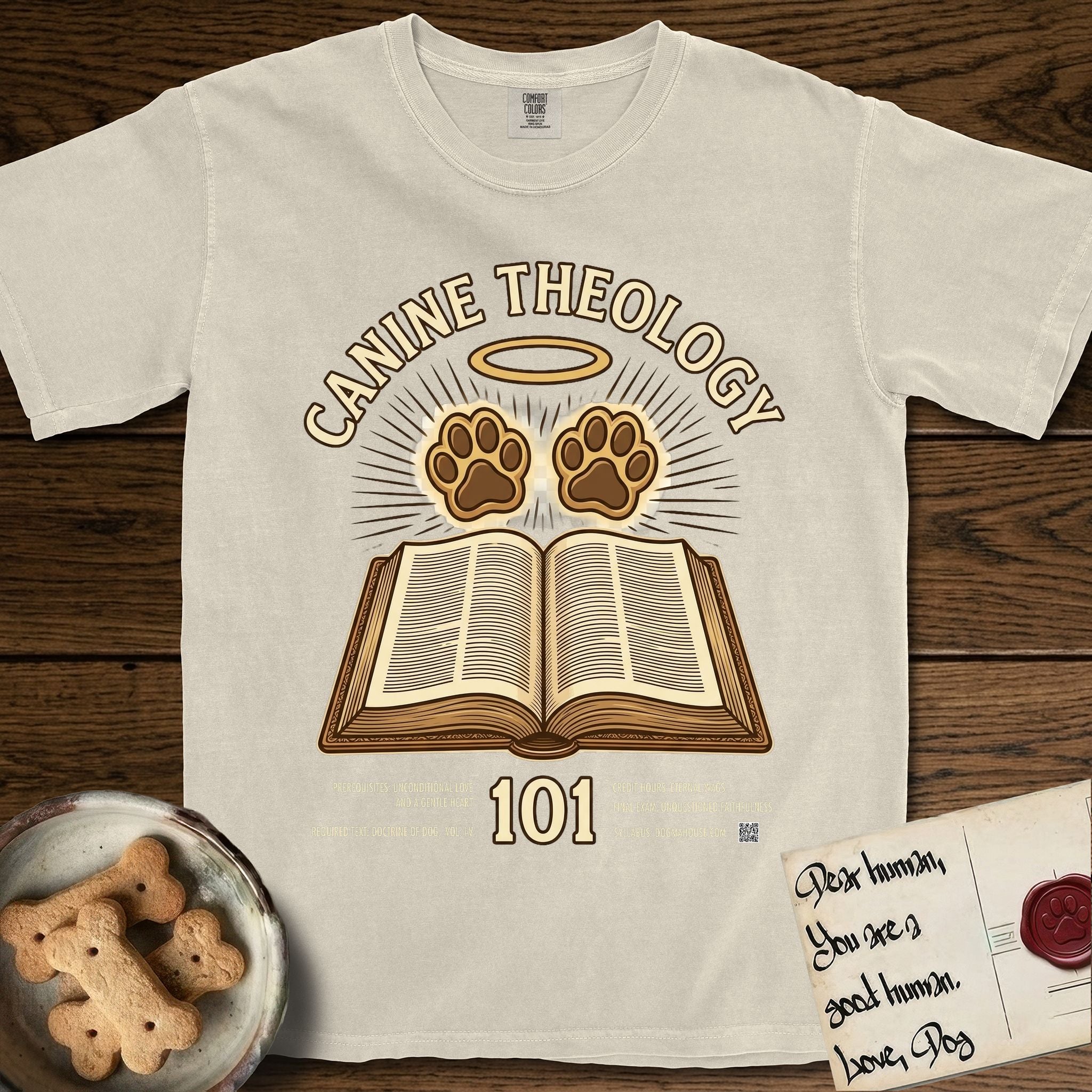 Canine Theology 101 T-Shirt flat lay in Ivory, featuring a cream badge with an open book and two glowing paw prints rising above it, ringed by a circular halo.
