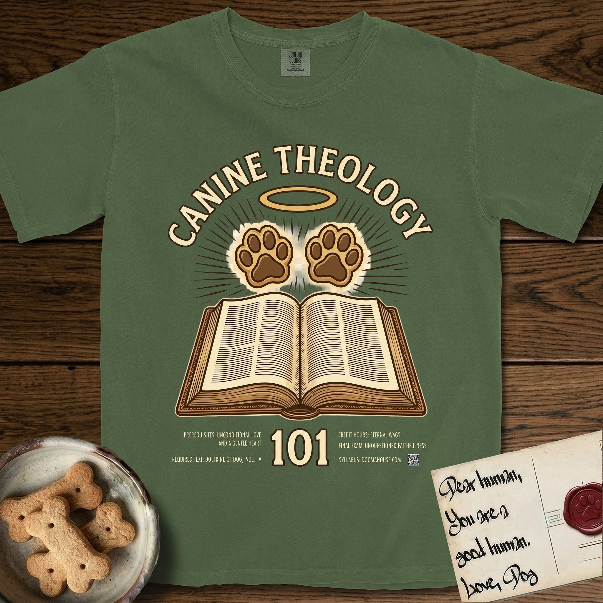 Canine Theology 101 T-Shirt flat lay in Moss, featuring a cream badge with an open book and two glowing paw prints rising above it, ringed by a circular halo.