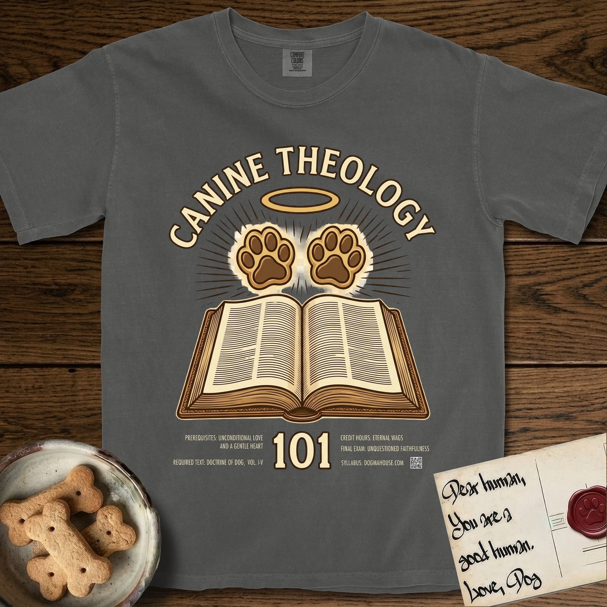 Canine Theology 101 T-Shirt flat lay in Pepper, featuring a cream badge with an open book and two glowing paw prints rising above it, ringed by a circular halo.