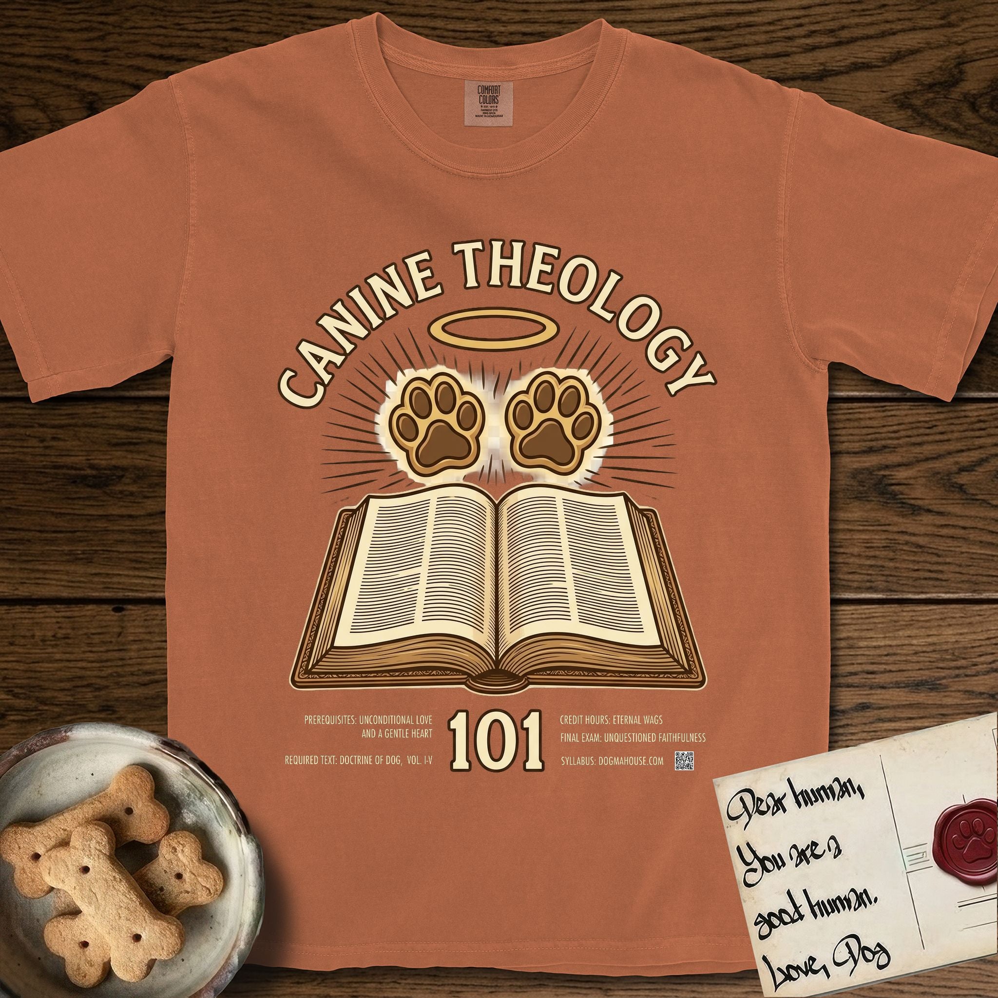 Canine Theology 101 T-Shirt flat lay in Yam, featuring a cream badge with an open book and two glowing paw prints rising above it, ringed by a circular halo.