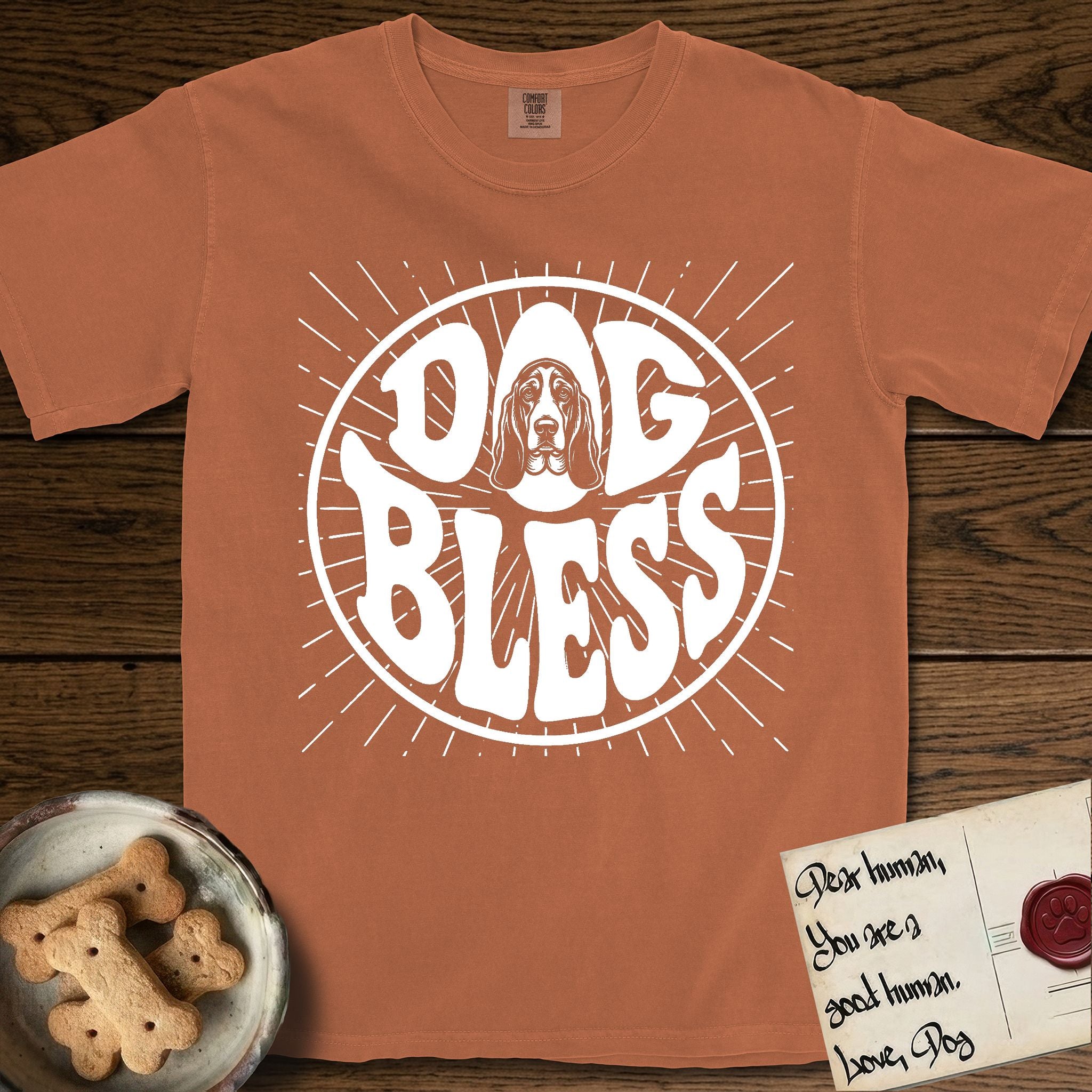 Dog Bless Basset Hound T-Shirt flat lay in Yam, featuring Retro bubble-letter badge reading DOG BLESS with a long-eared Basset Hound face framed in the letter O, hand-drawn sunburst rays radiating outward.