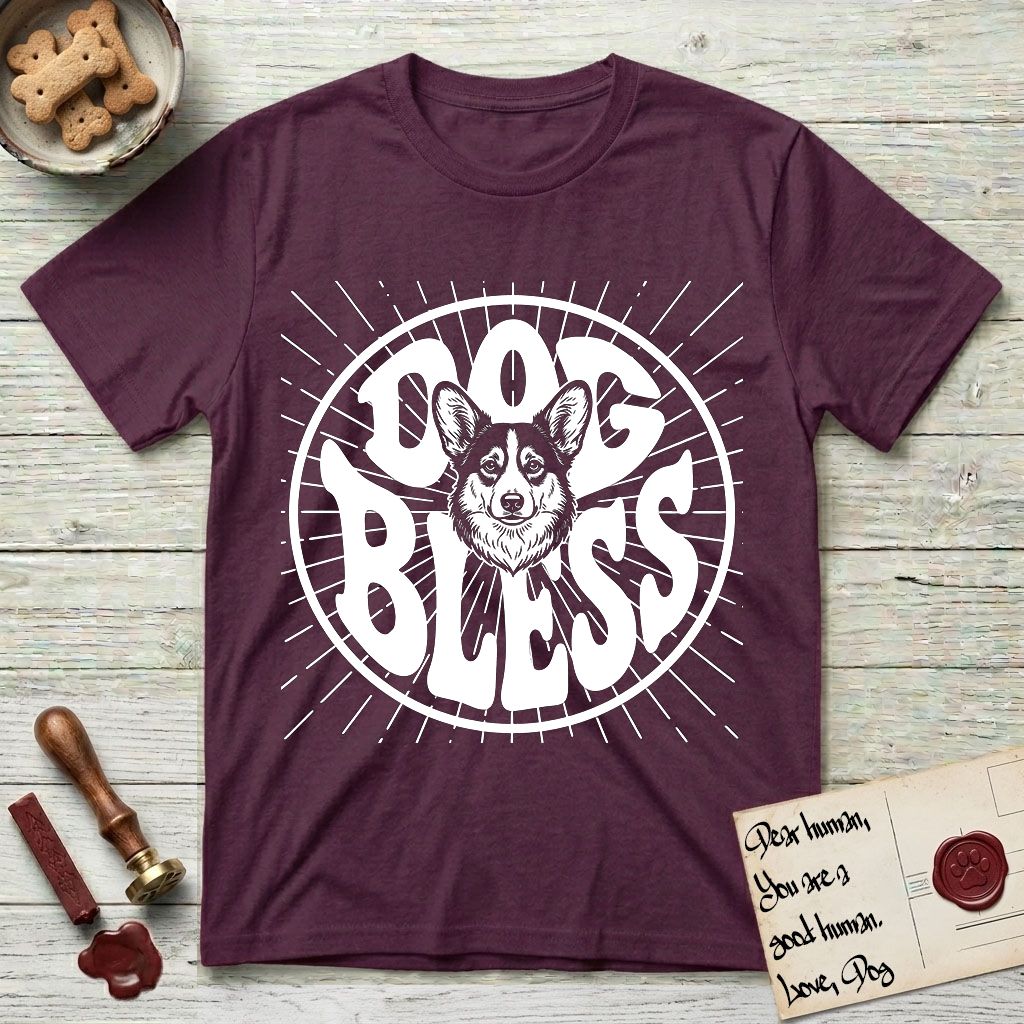 Dog Bless Corgi T-Shirt flat lay in Maroon, featuring Retro bubble-letter badge reading DOG BLESS with a smiling Corgi face framed in the letter O, chest fluff spilling below the letter, hand-drawn sunburst rays radiating outward.