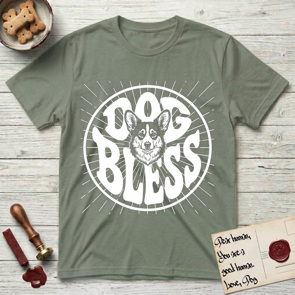 Dog Bless Corgi T-Shirt flat lay in Sage, featuring Retro bubble-letter badge reading DOG BLESS with a smiling Corgi face framed in the letter O, chest fluff spilling below the letter, hand-drawn sunburst rays radiating outward.