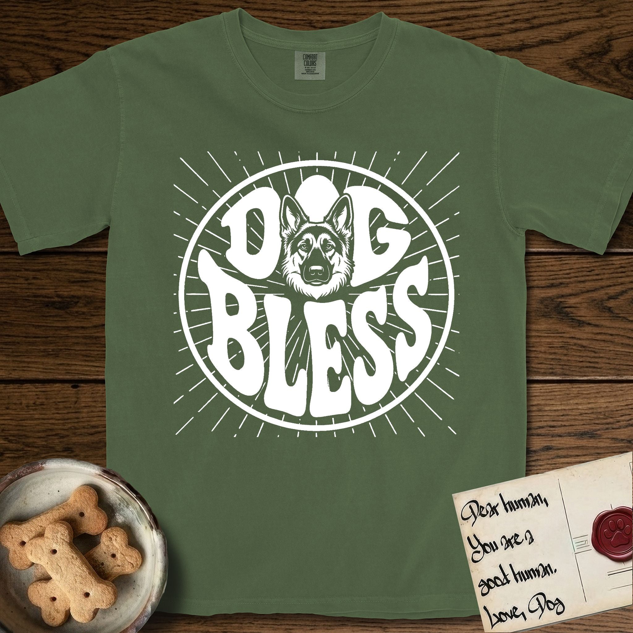 Dog Bless German Shepherd T-Shirt flat lay in Moss, featuring Retro bubble-letter badge reading DOG BLESS with a German Shepherd face with tall upright ears framed in the letter O, hand-drawn sunburst rays radiating outward.